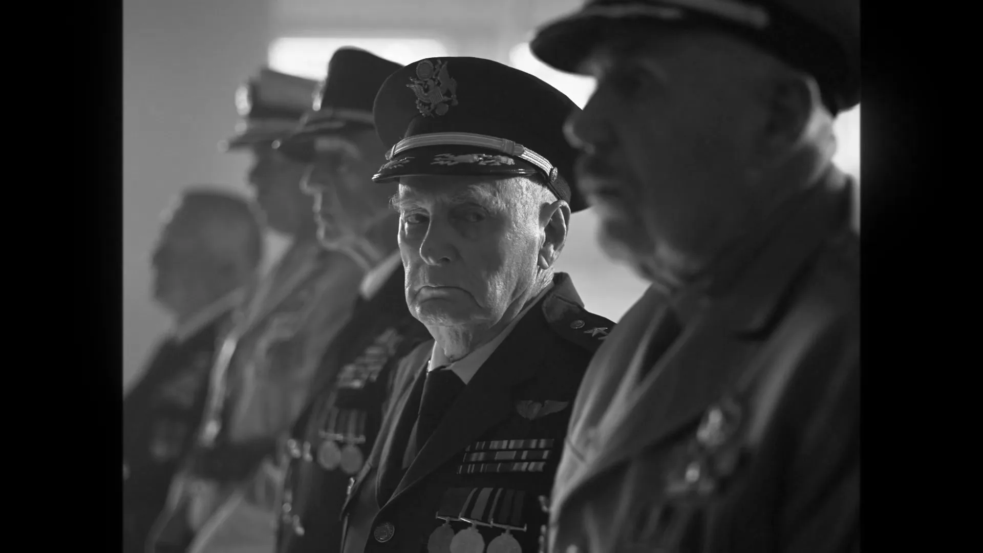 A group of older men in military uniforms and hats stand in a line, looking forward with serious expressions in a black and white shot.