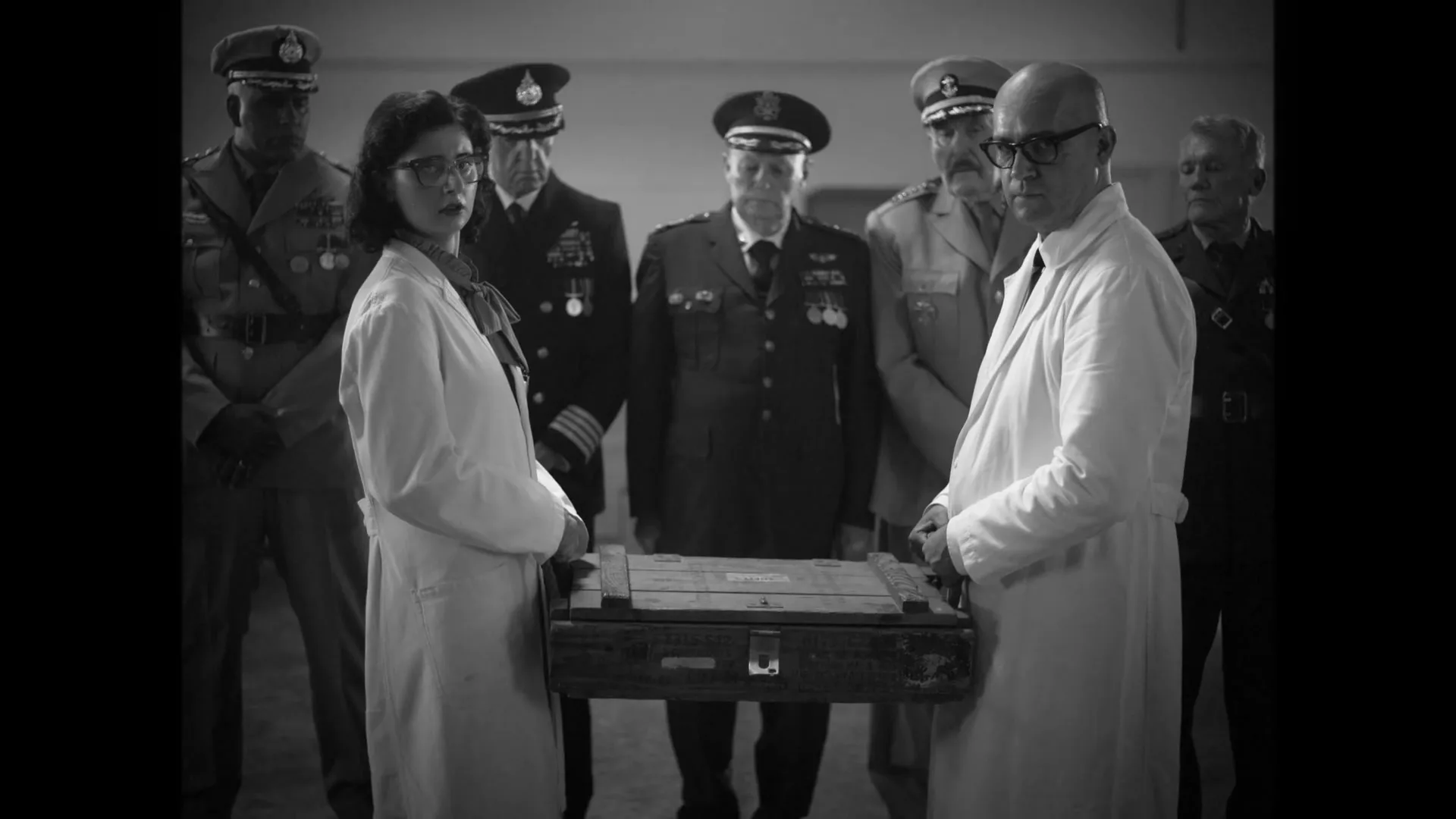 A group of military officers and scientists observe a wooden box in a large, dimly lit hangar, in black and white.