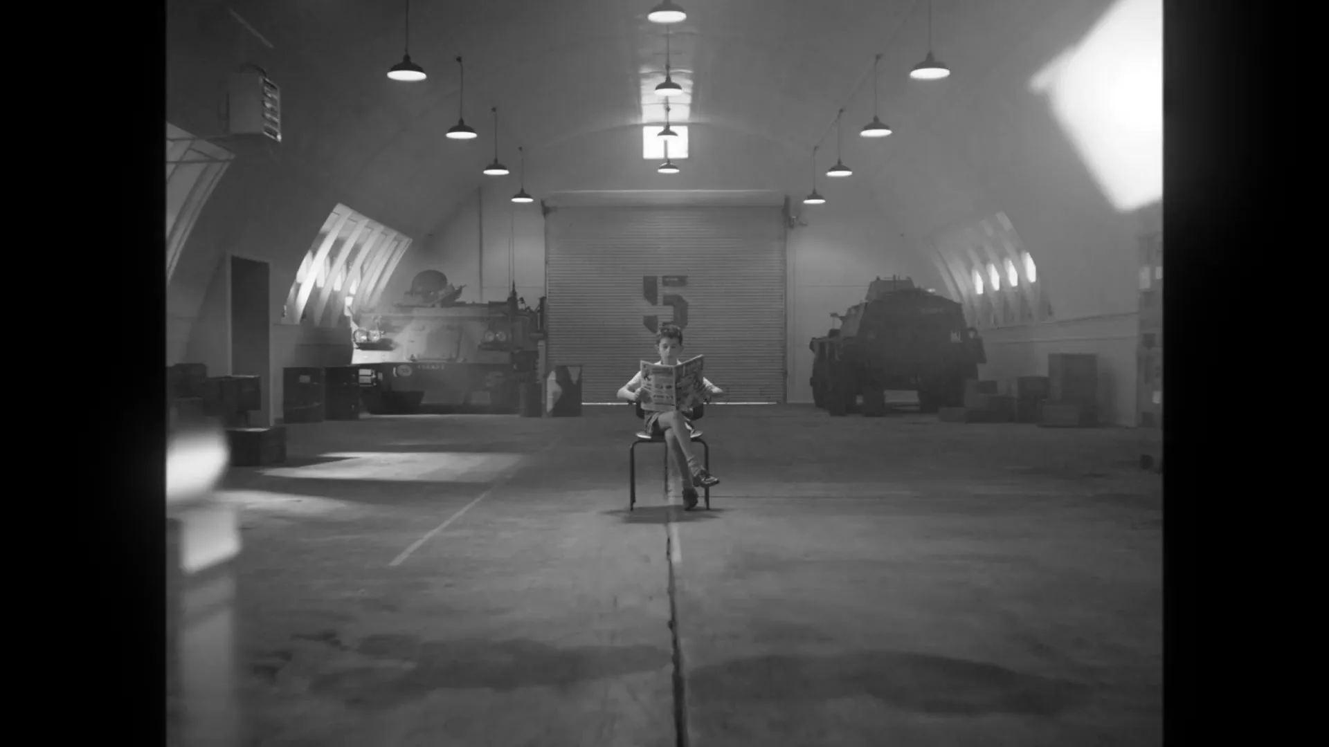 A young boy, sitting on a chair in a large hangar, is covered in various metal objects as he reads a newspaper.