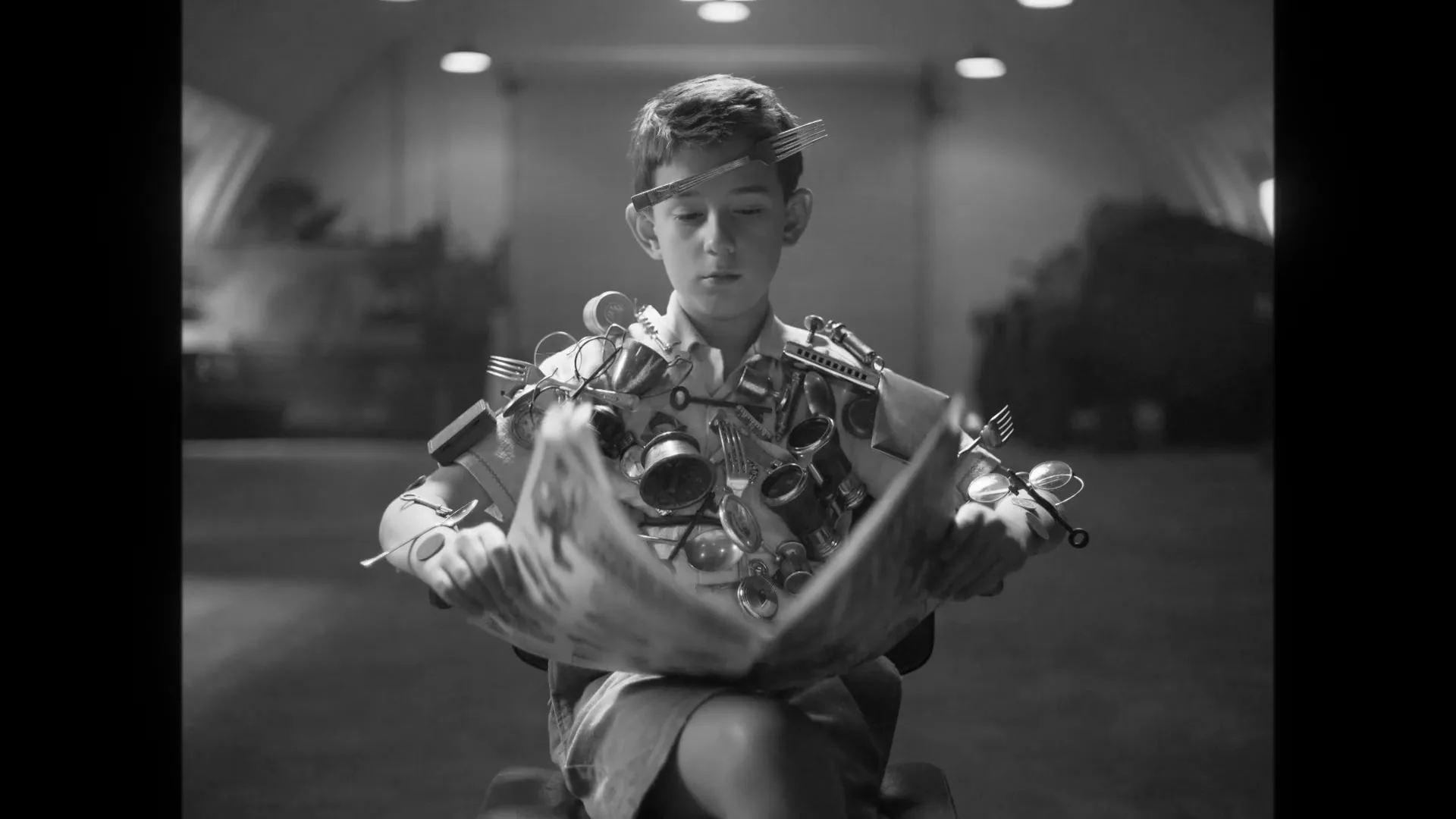 A young boy, covered in metal objects, sits in a chair as a group of military officers applaud him in a large hangar.