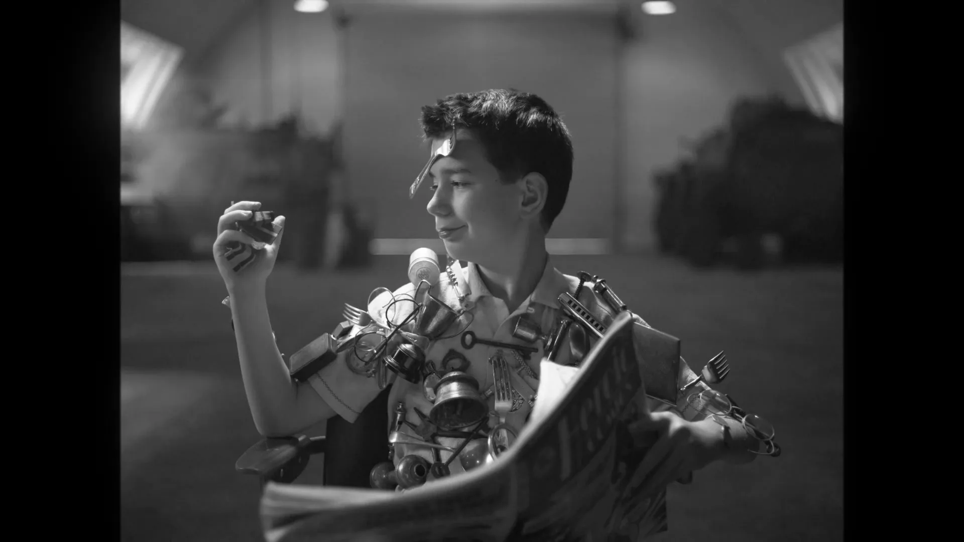 A young boy, covered in metal objects, sits on a chair in a hangar, reading a newspaper and looking at a small metal object in his hand.