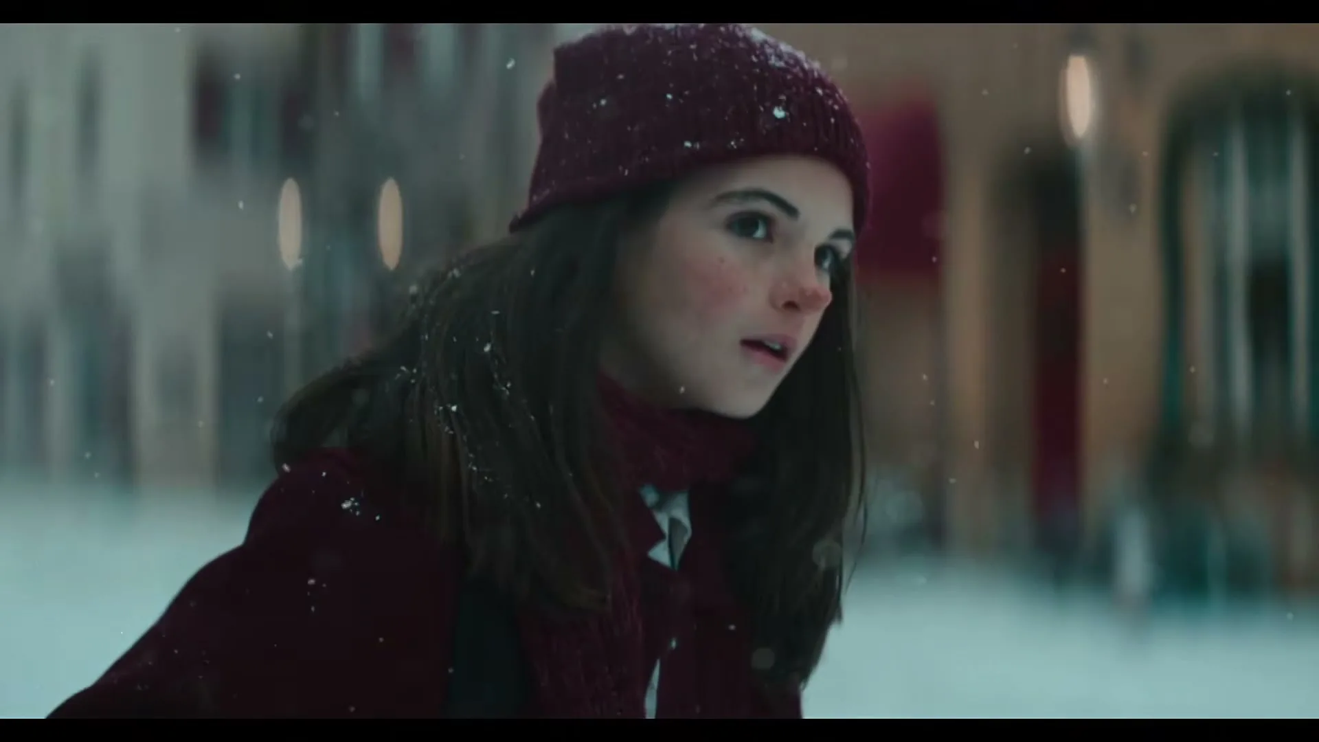 A young girl with dark hair and a maroon hat and scarf looks around while ice skating outdoors as snow falls.