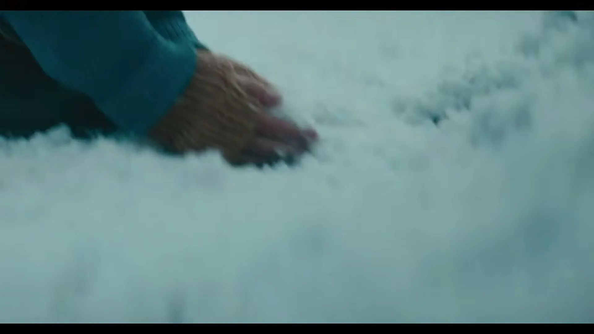 A person in a blue sweater and brown gloves kneels in the snow, forming a snowball with their hands.