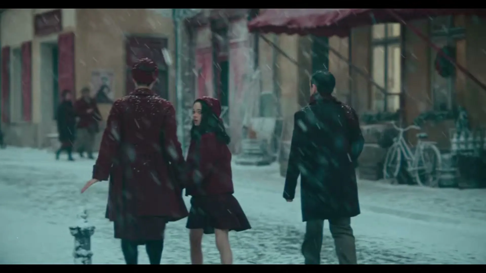 A family of three, dressed in dark coats, walks away from the camera down a snowy street in a European town as snow falls.