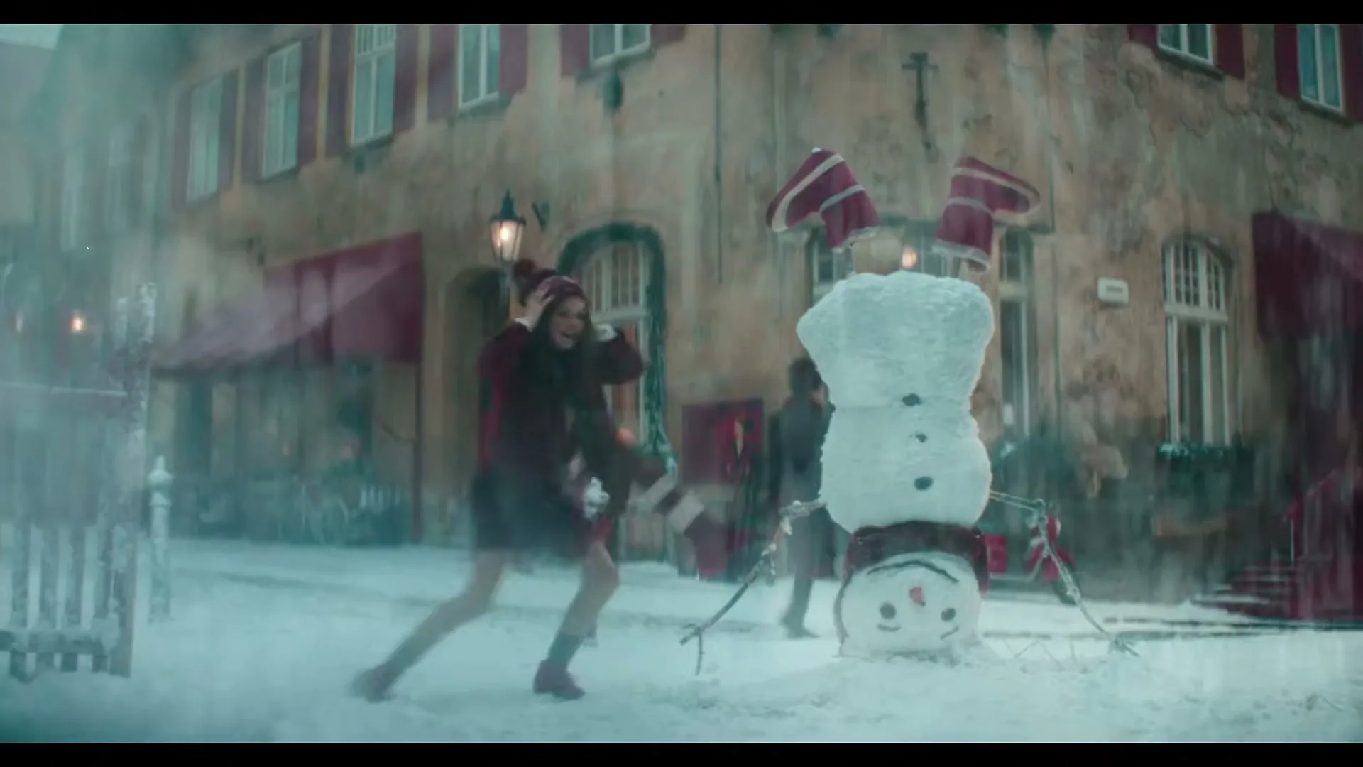 A young woman in winter clothes dances playfully in a snowy town square next to an upside-down snowman, as snow falls around her.