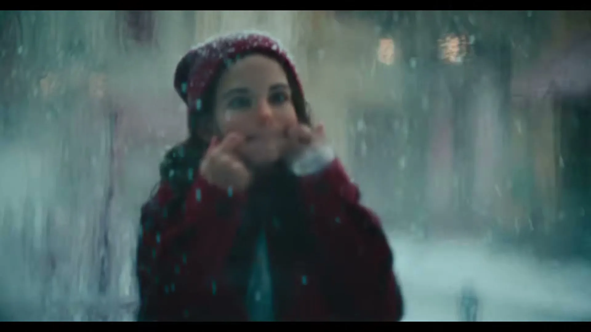 A young girl in a red hat and coat makes a funny face through a rainy window, with snow falling outside on a winter day.