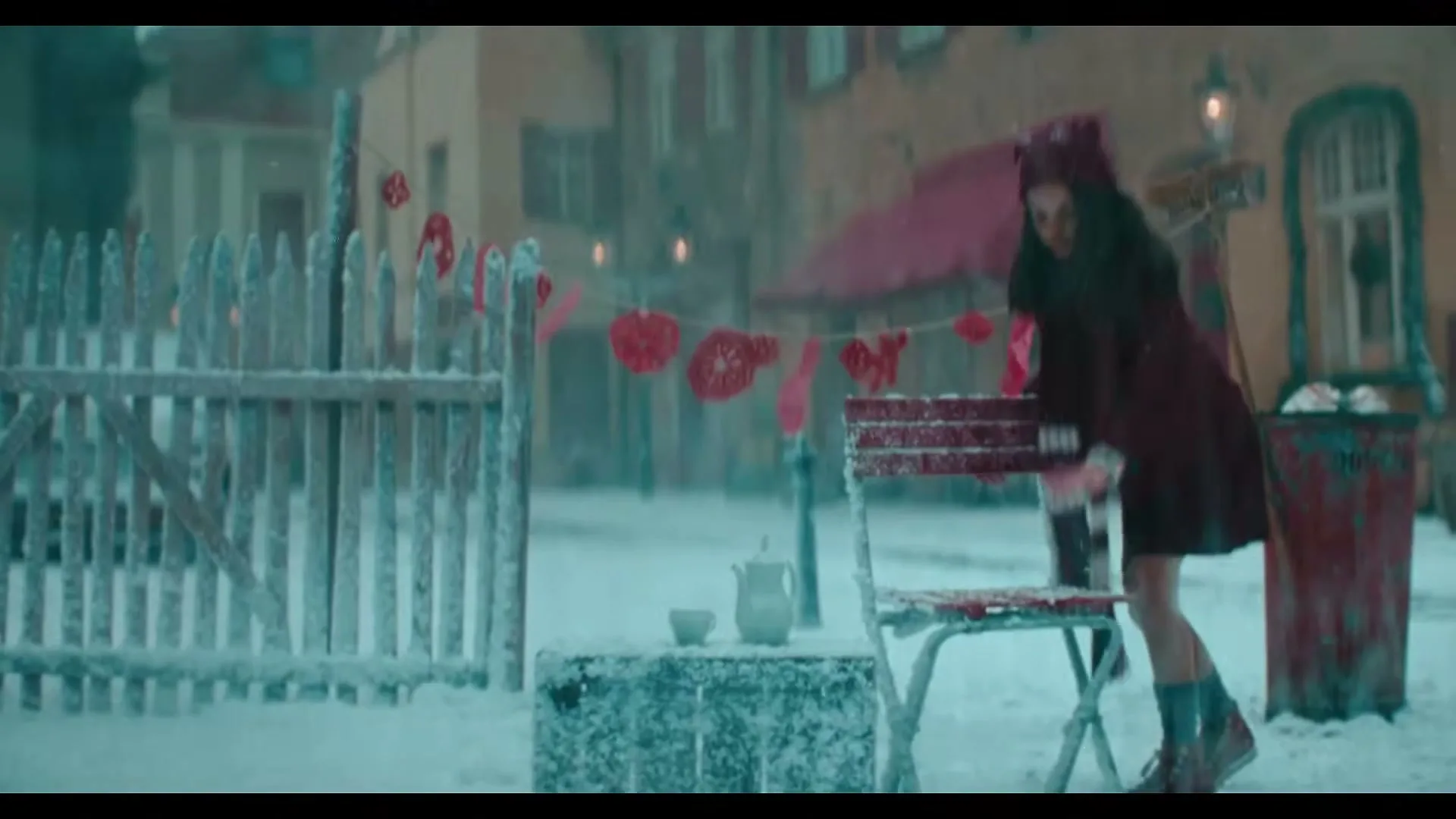 A young woman in a red coat and hat wipes snow off a red chair in a snowy, festive European town square.