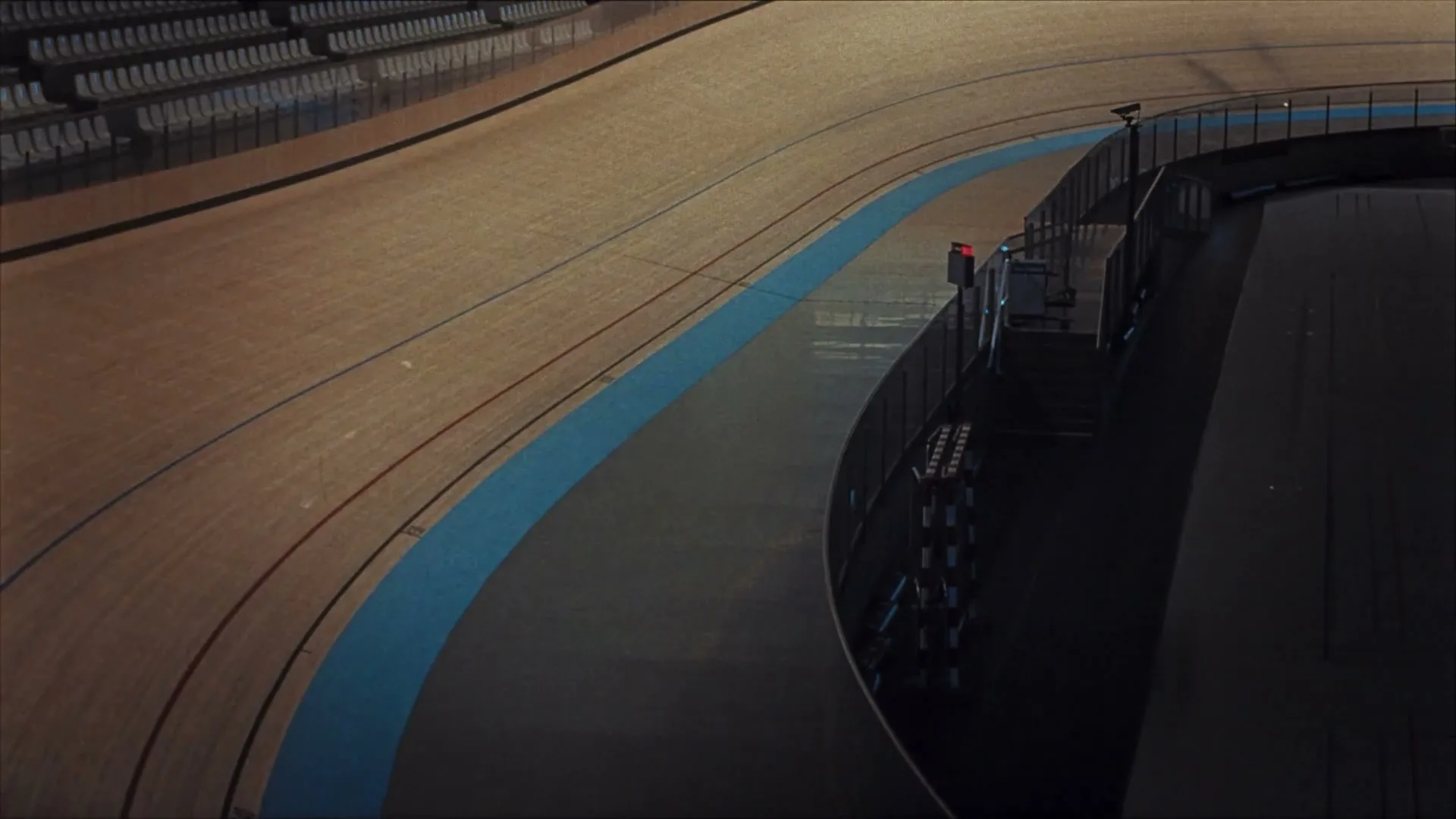 An empty velodrome track with spectator seating in the background is slowly illuminated by a rising light source.