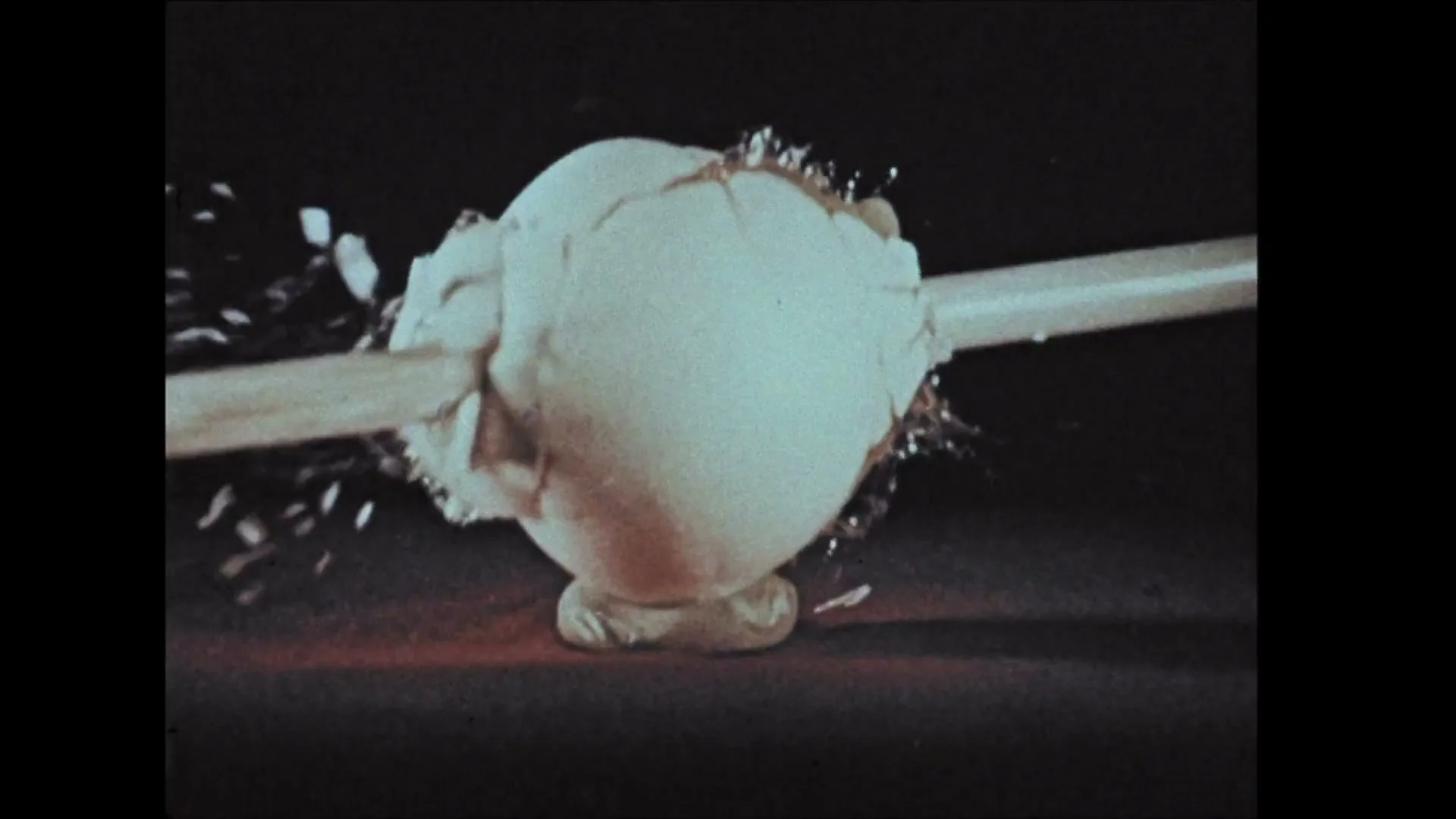 A white egg is hit by a projectile, shattering and splattering its contents in slow motion against a dark background.