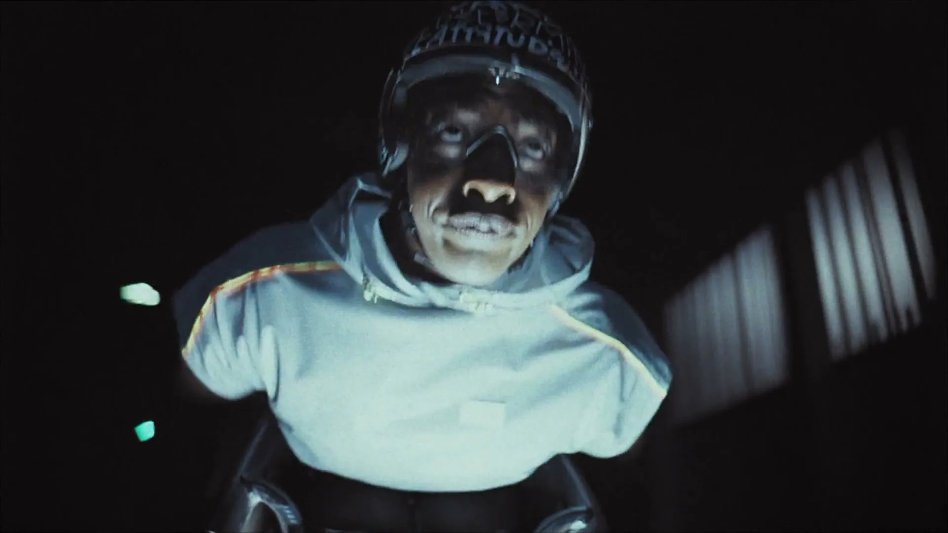 A male para-athlete in a helmet and hoodie, with a determined expression, speeds forward in a dark, enclosed space.