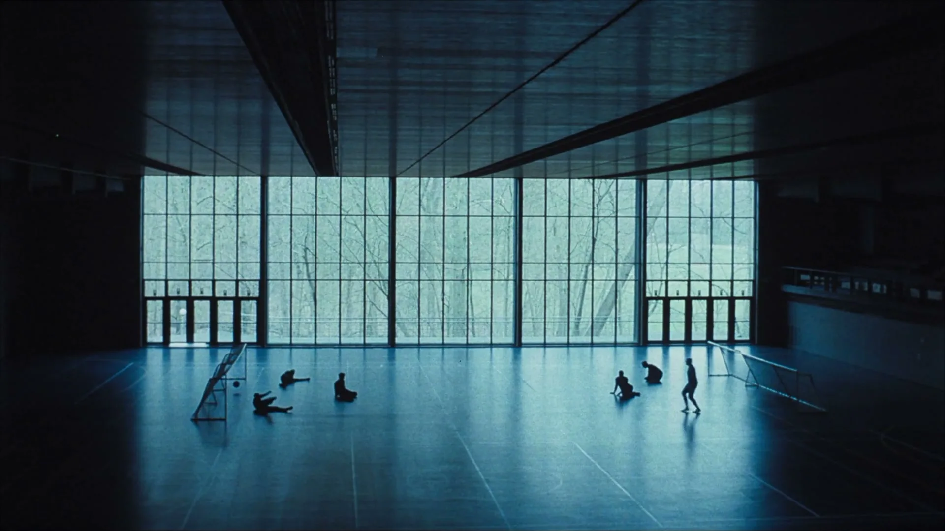 Silhouetted athletes with disabilities play a game of adapted football on a polished gym floor, illuminated by a large window overlooking a forest.