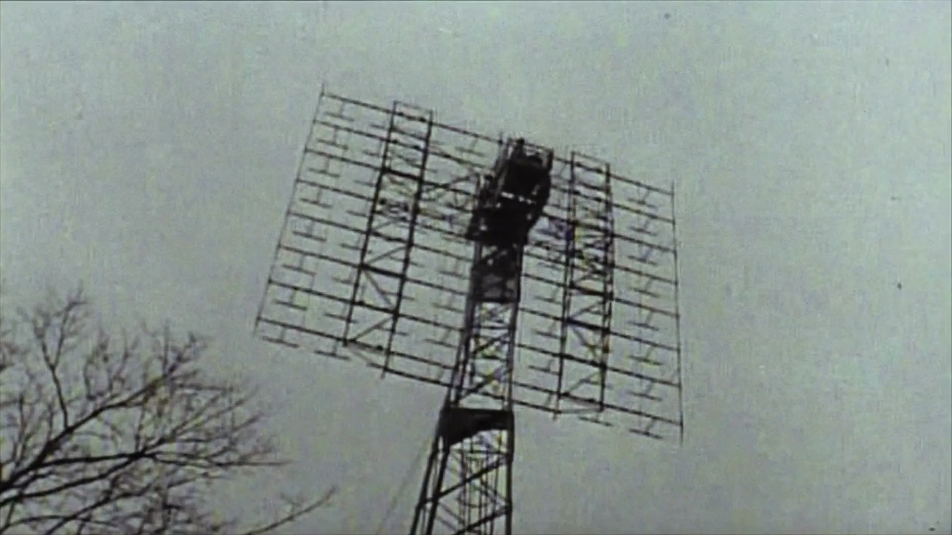 A large, square radar dish rotates slowly against a grey sky, with bare tree branches visible in the lower left corner.