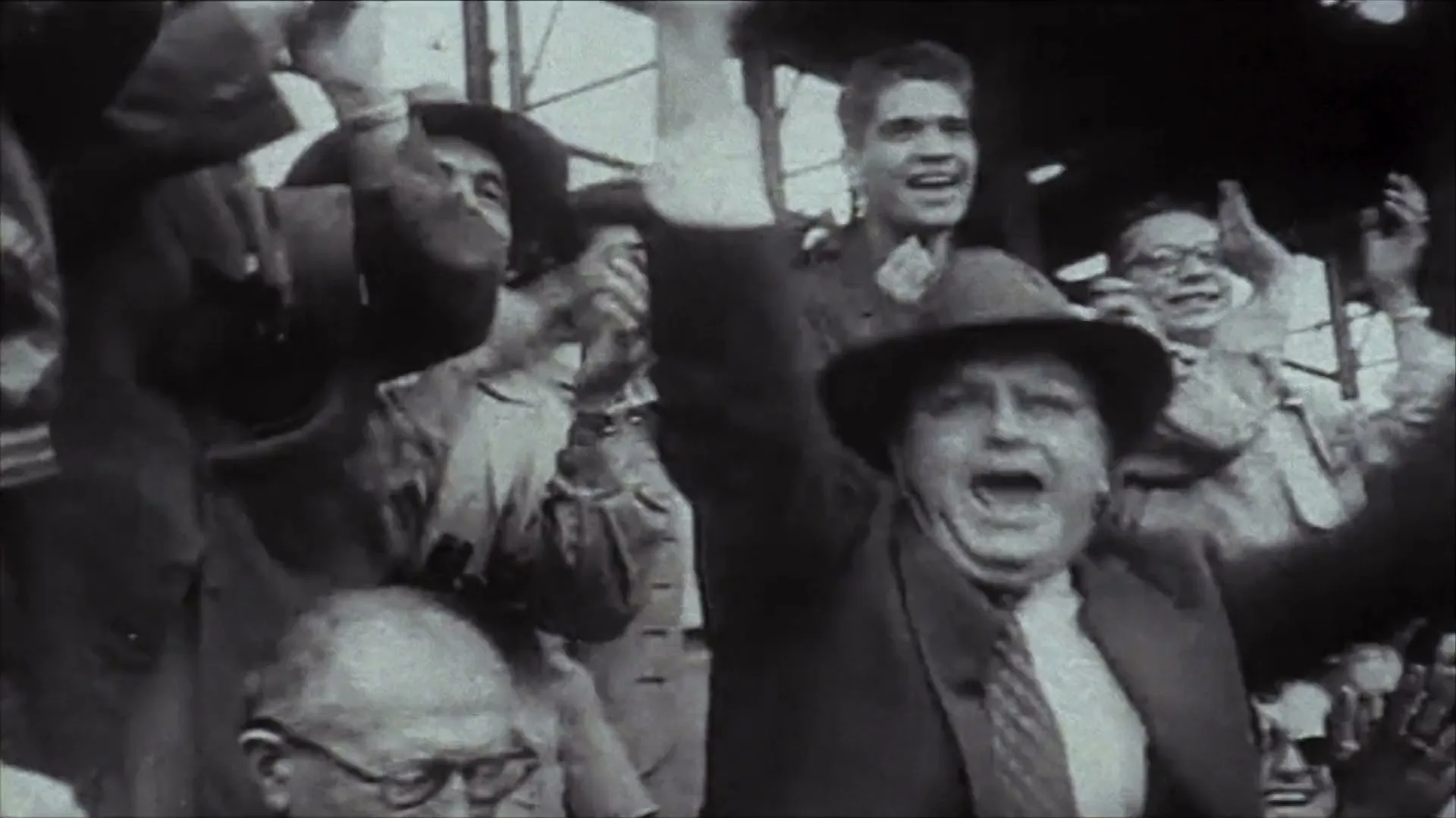 A crowd of people, mostly men, cheer excitedly with their arms raised in a black and white archival shot at a sporting event.