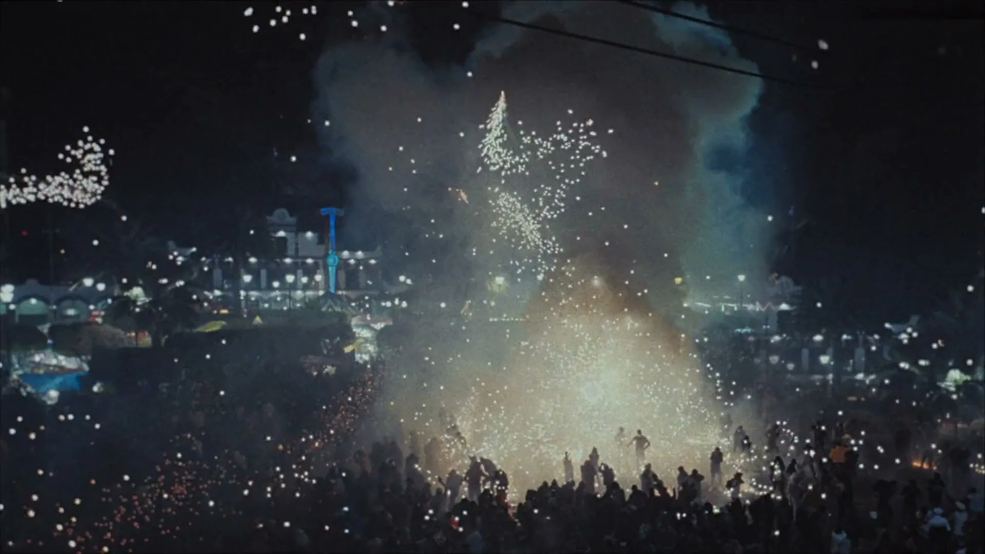 A large crowd of people are gathered at night, watching a fireworks display with bright sparks and smoke filling the air.