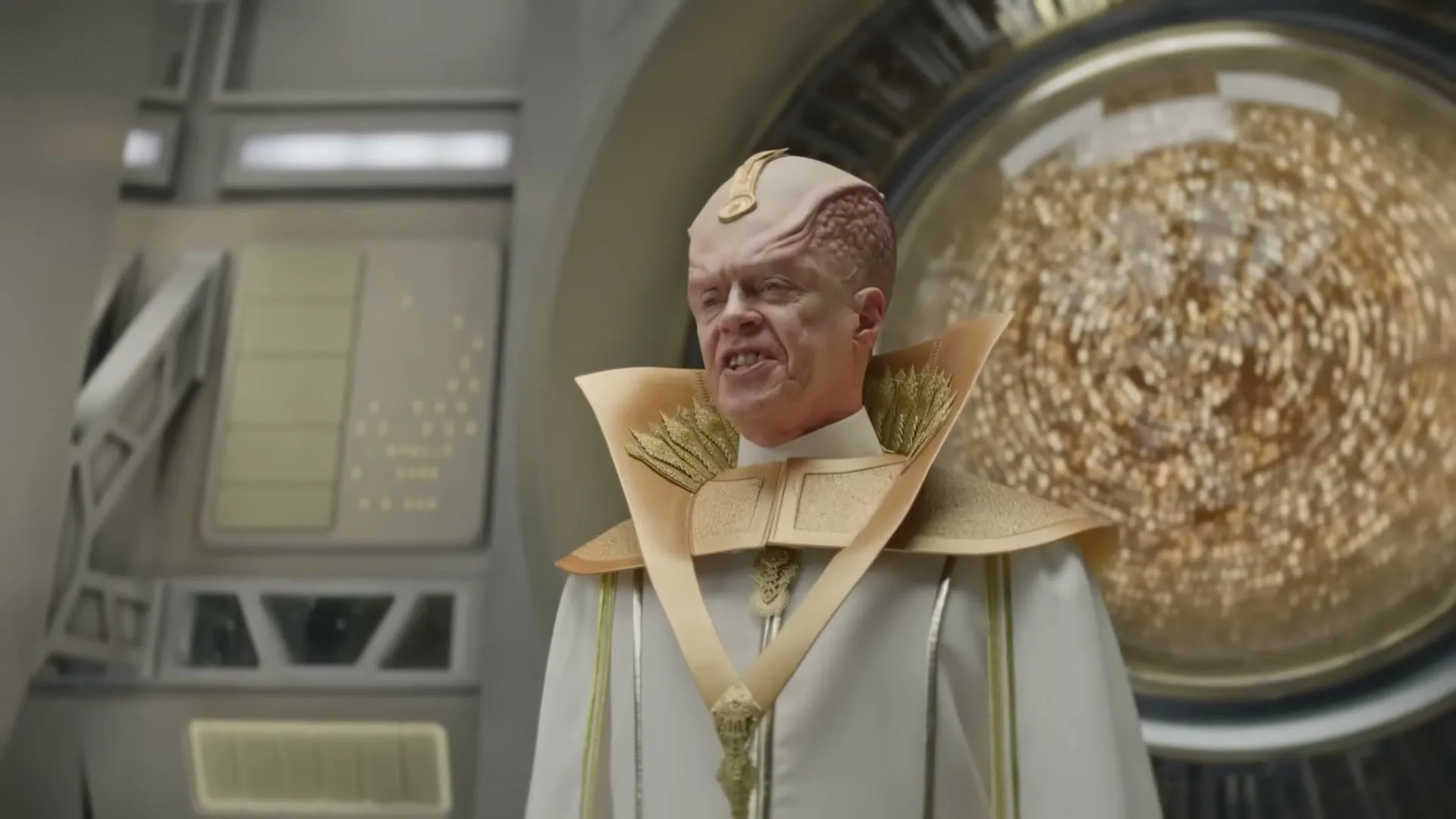 An alien leader, with pink skin and a gold and white uniform, stands in a futuristic control room, speaking with a determined expression.