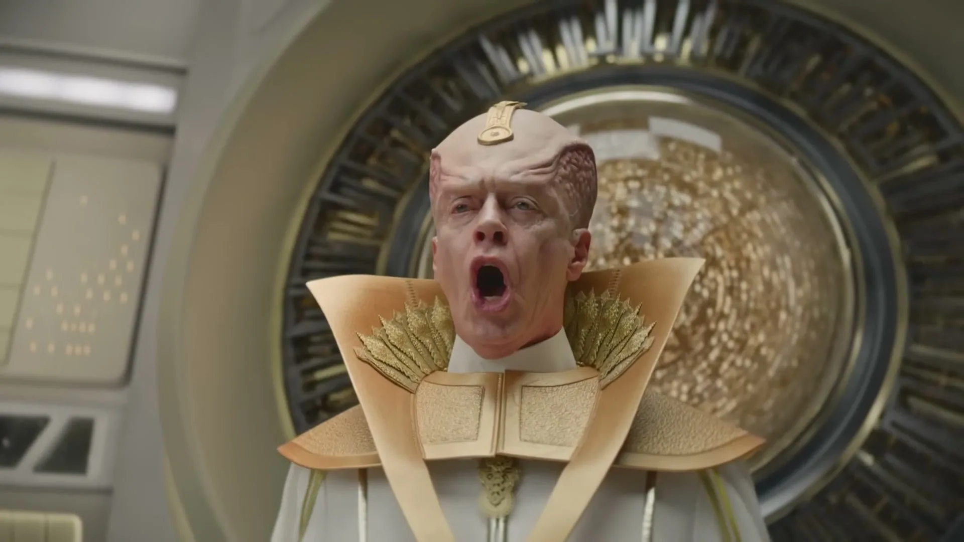 A male alien with a large, ridged head and golden robes shouts dramatically in a futuristic, metallic room with a large golden sphere in the background.
