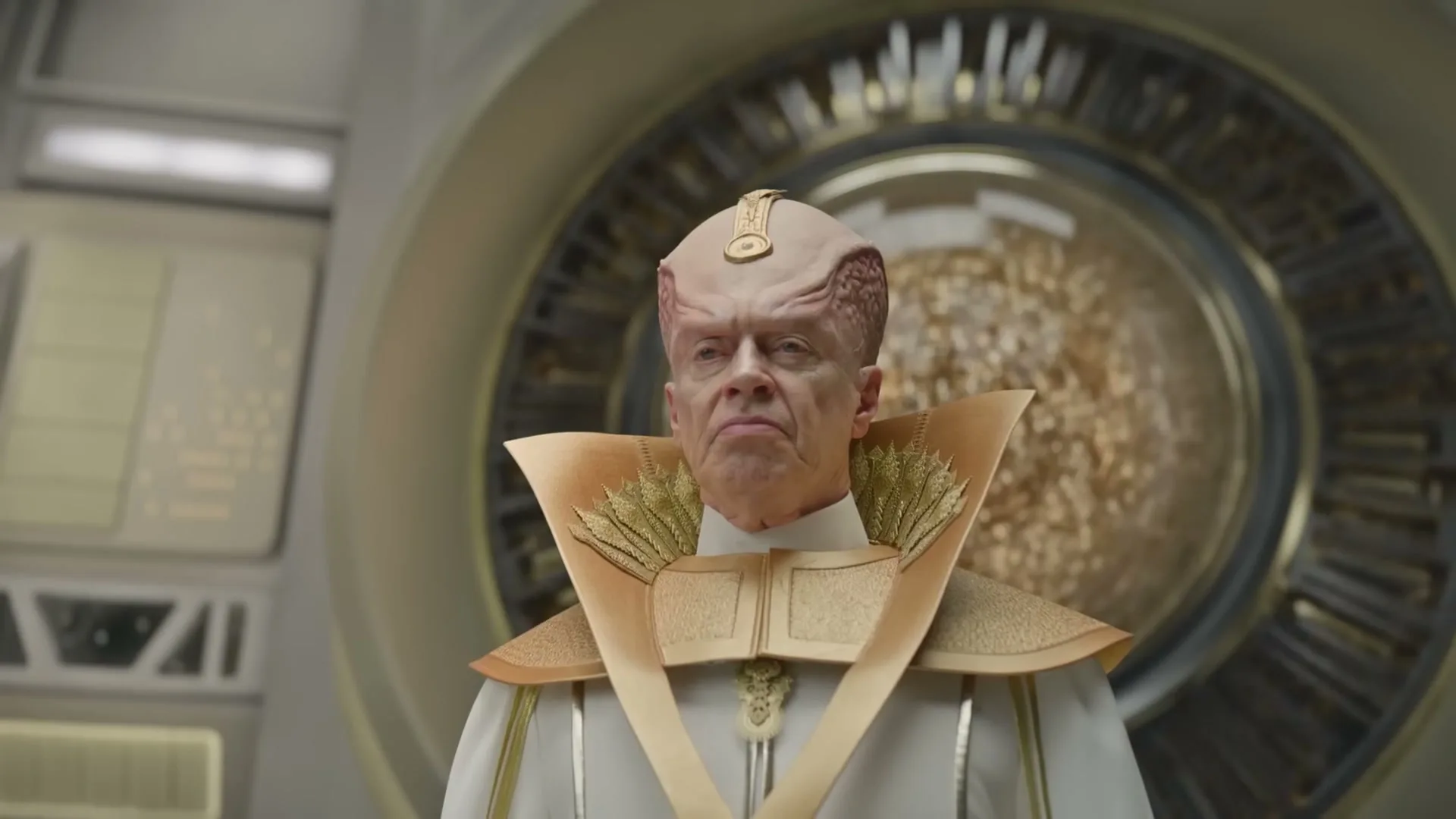 A male alien with a wrinkled, pink head and golden attire looks sternly at the camera in a futuristic, metallic room.