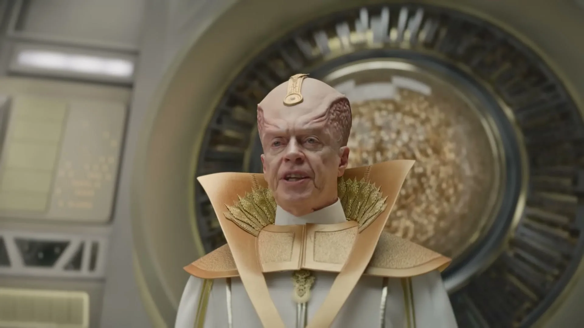 A male alien with a large brain-like head and golden robes looks annoyed in a futuristic control room.