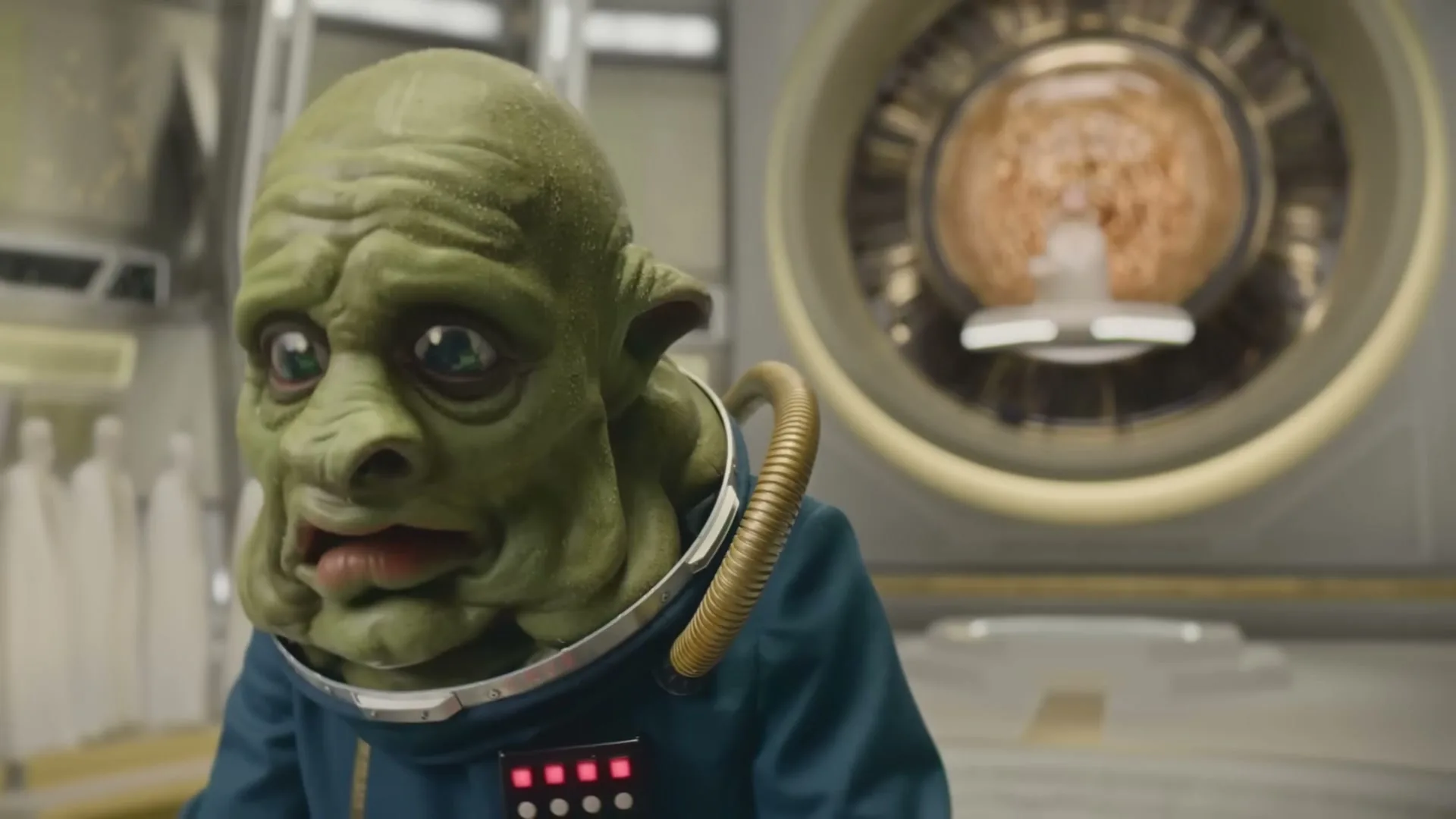 A green-skinned alien with large eyes and a blue uniform looks worried in a futuristic, sterile room with a glowing orb in the background.