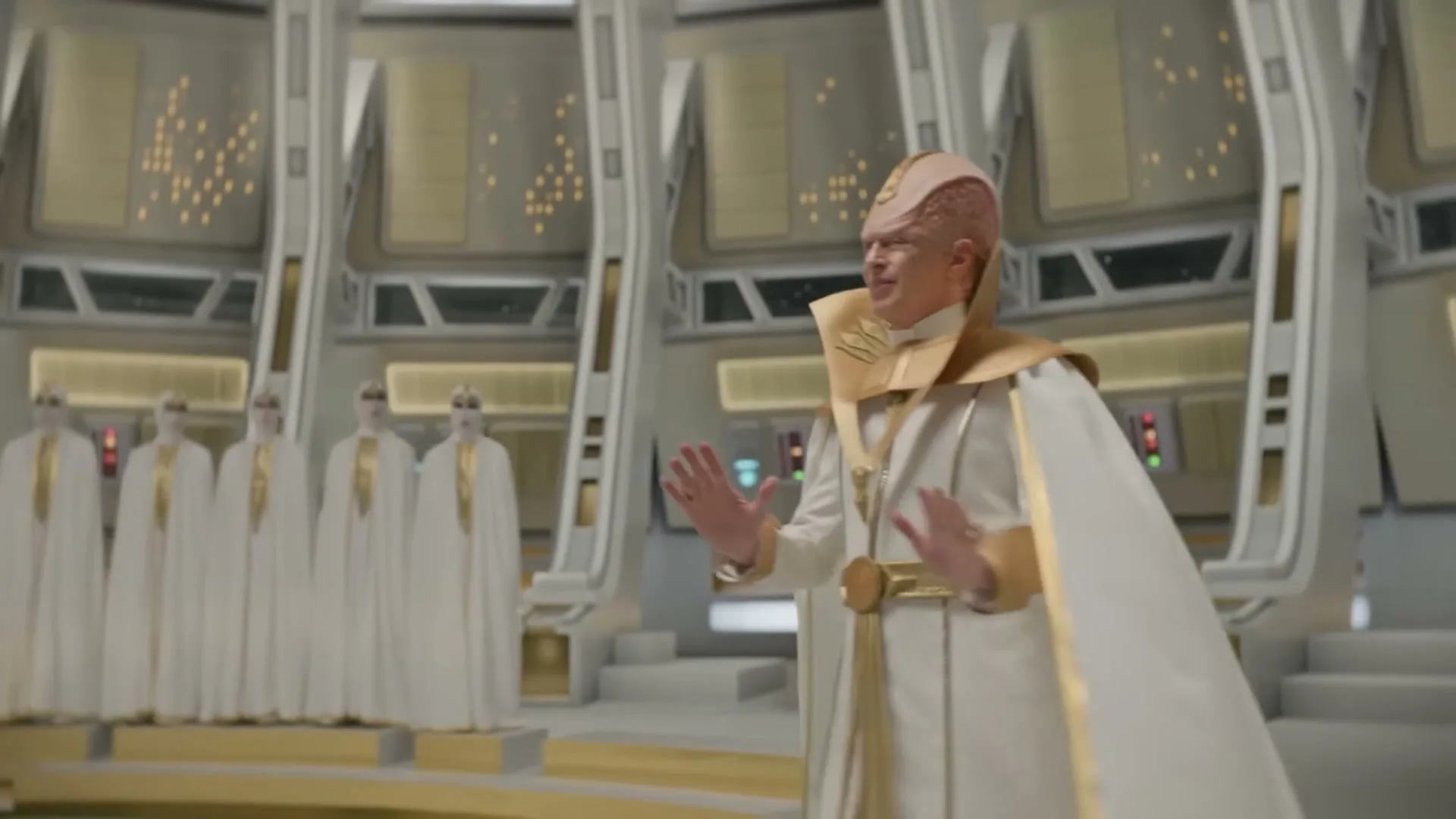 A bald, pink-skinned alien in a white and gold robe walks through a futuristic, white and gold chamber, gesturing with his hands.