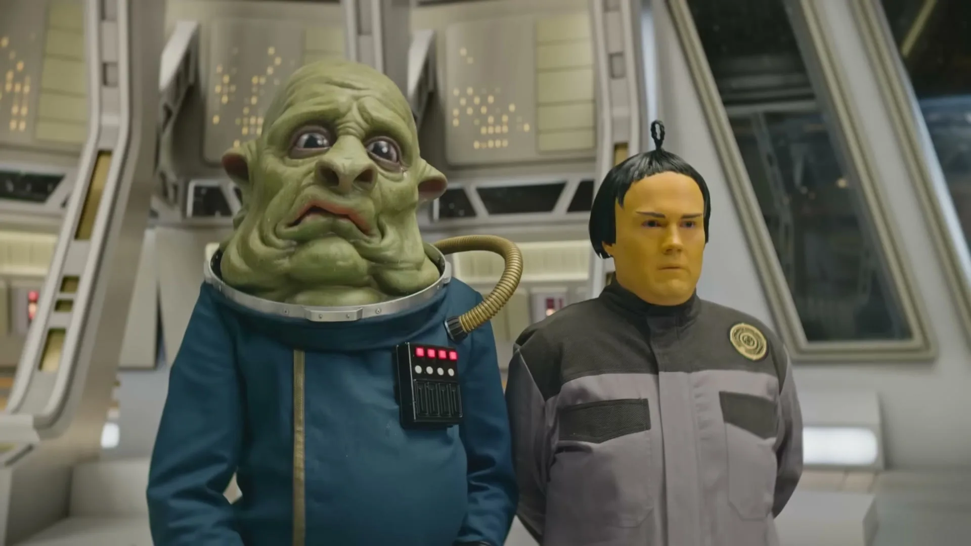 Two alien-like figures, one green and one yellow-faced, stand side-by-side in a futuristic, sterile interior setting, looking forward with neutral expressions.
