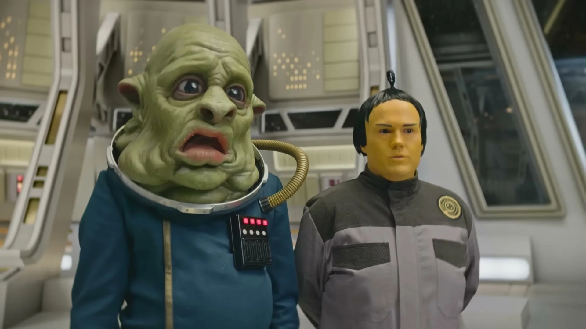 Two aliens, one green and one yellow, stand side-by-side in a futuristic spaceship interior, looking concerned.