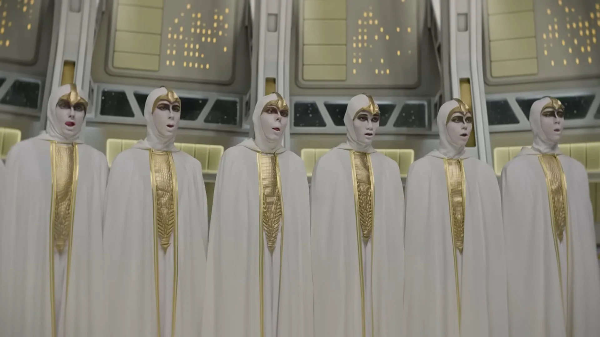 Six people in white hooded robes with gold accents and white face paint stand in a futuristic room with glowing panels.