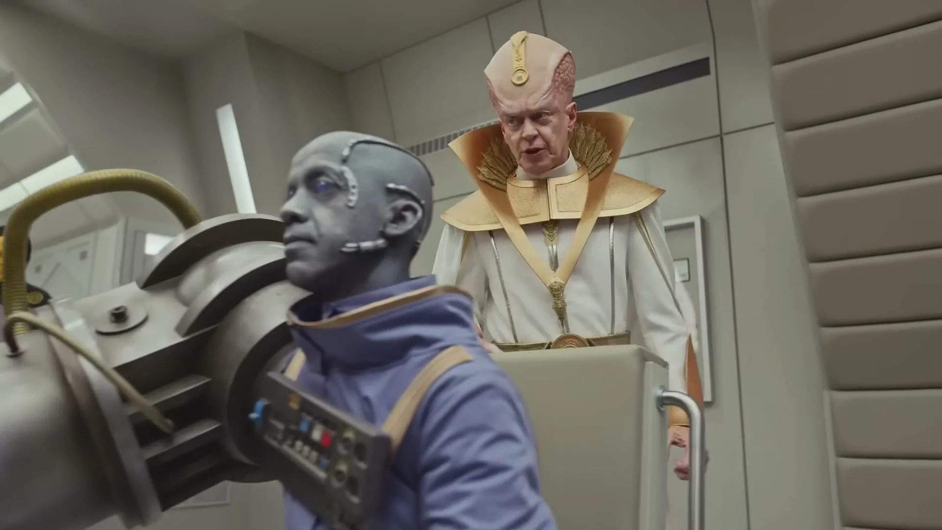 A blue-skinned alien in a purple suit sits at a control panel, while a bald, pale-skinned alien in a white and gold robe stands behind him, urging him on in a futuristic room.