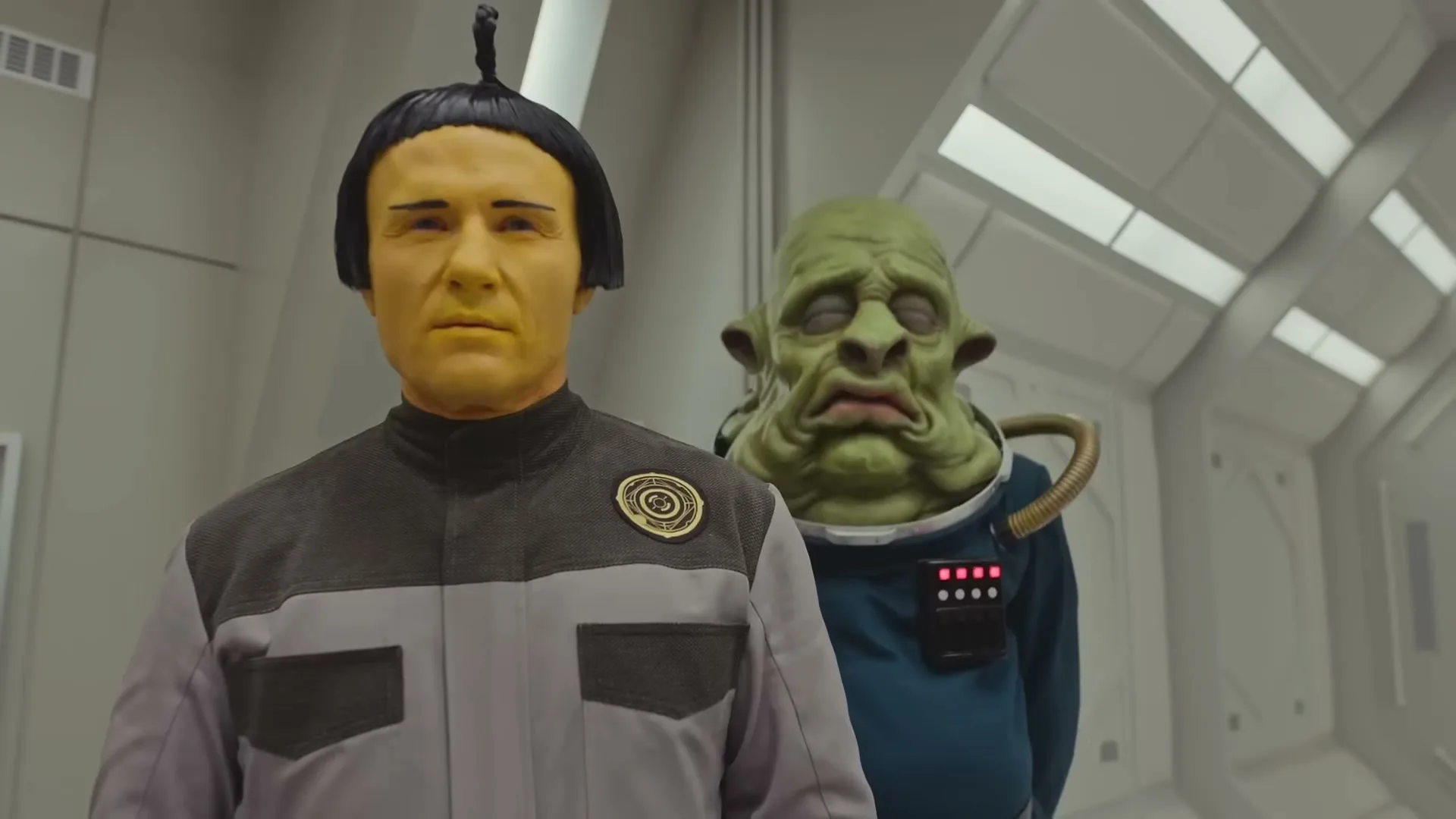 A yellow-faced man in a grey uniform stands in a spaceship corridor, with a green alien looking over his shoulder.