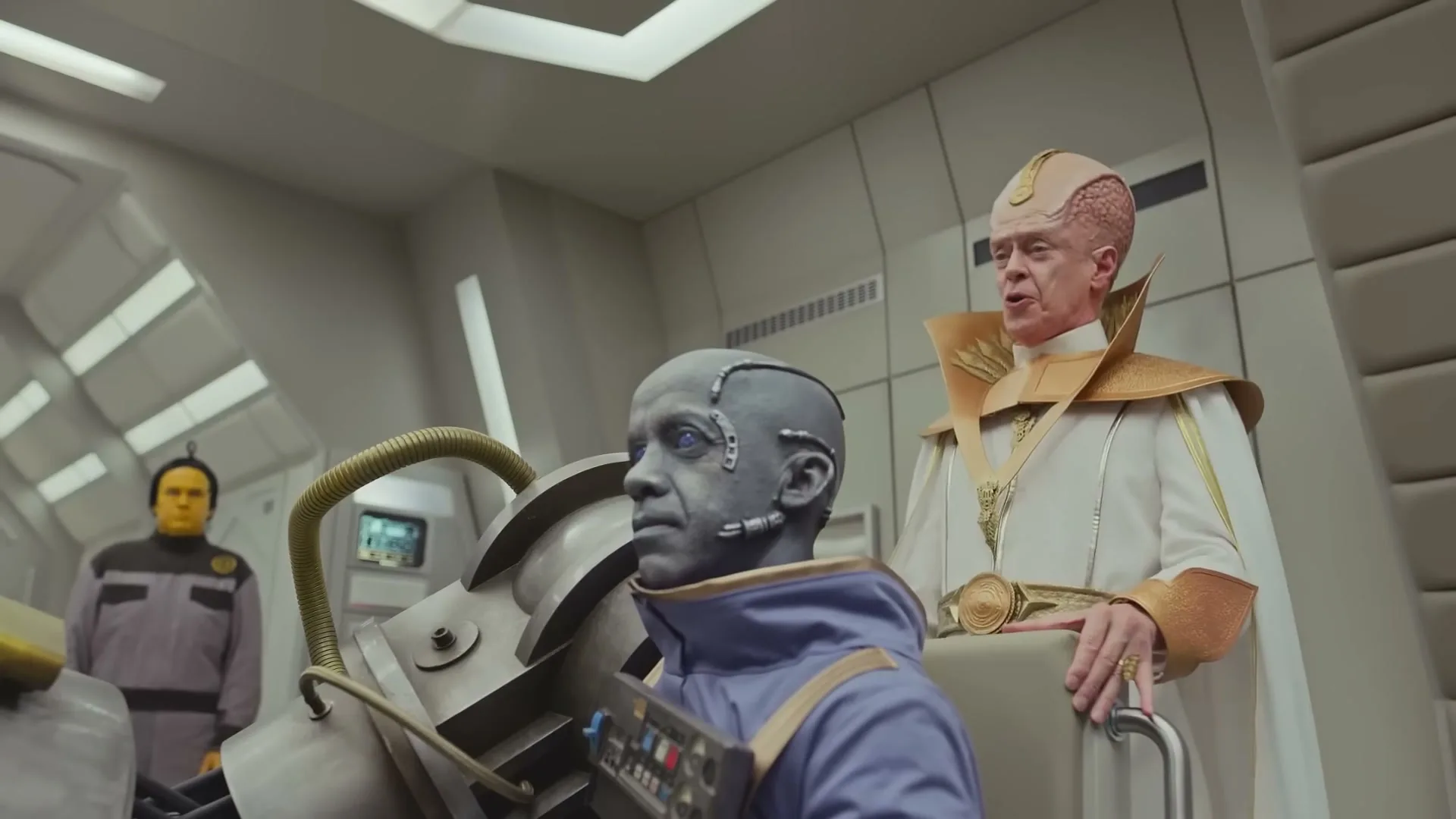 A man with a bald, pink, ridged head and a white and gold robe stands next to a blue-skinned man in a futuristic chair, in a spaceship control room.