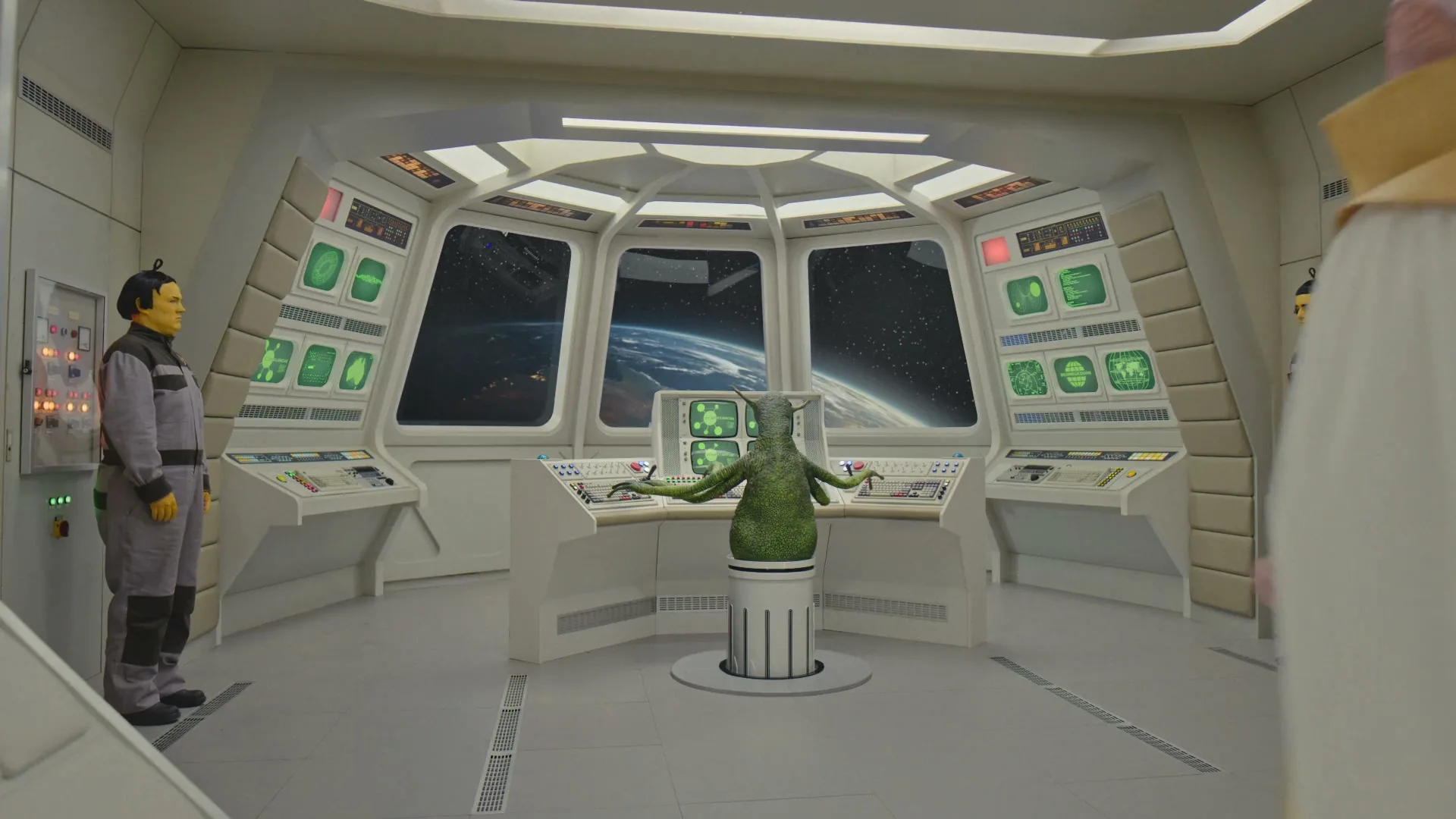 A green alien sits at a control panel in a spaceship, flanked by two humanoids, as a person in a white gown enters the frame.