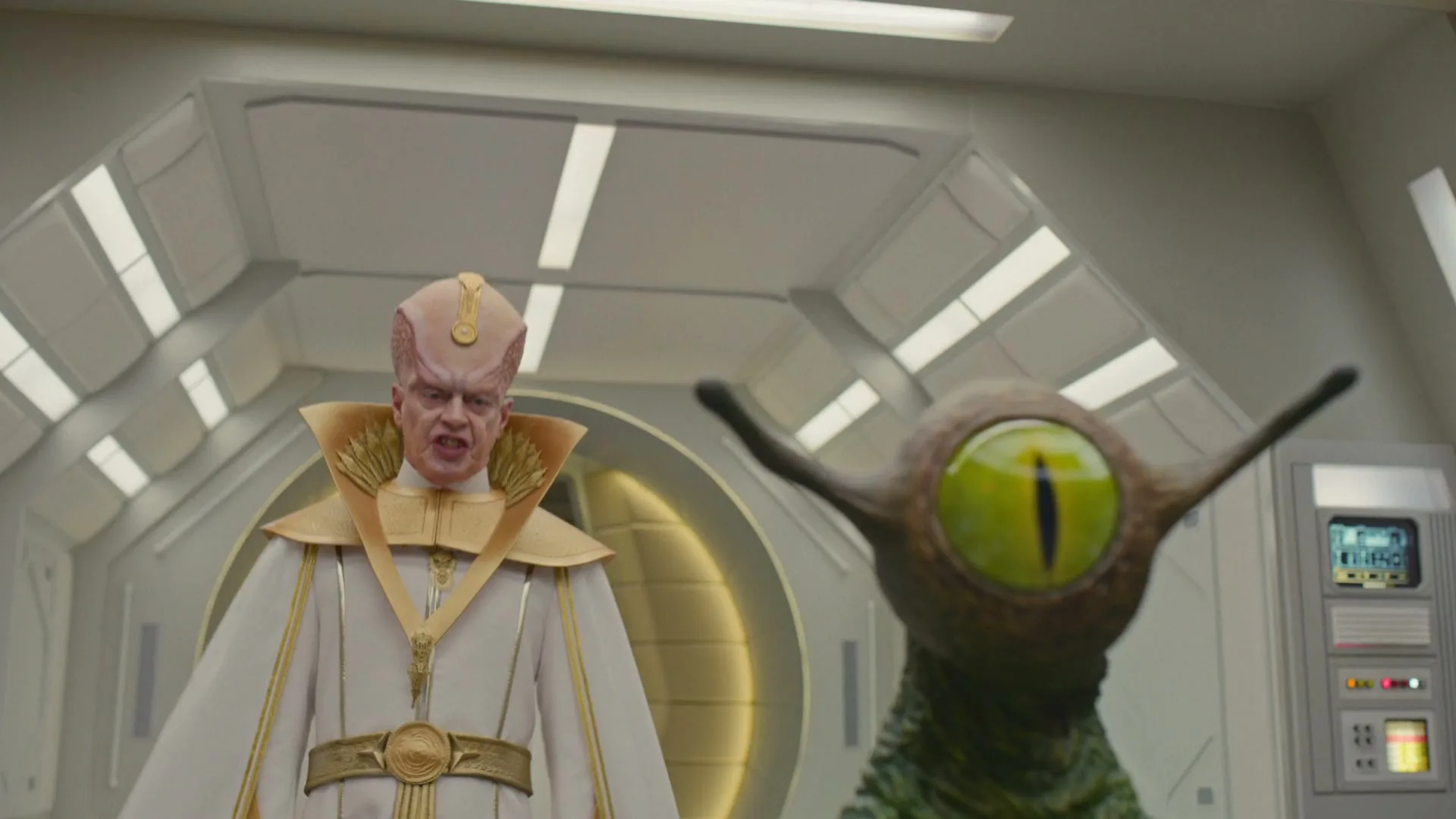 A male alien in a white and gold robe stands next to a green alien with a large eye in a futuristic spaceship interior.