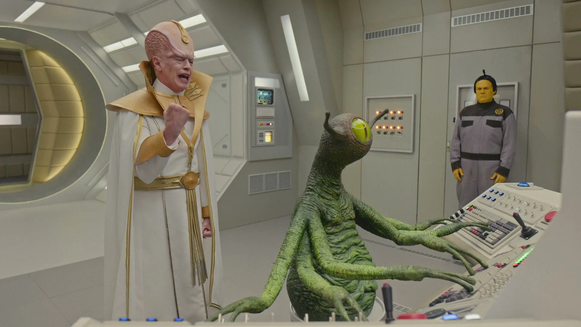 A male alien in a white and gold robe stands next to a green alien with multiple arms at a control panel, while another yellow-skinned alien stands in the background on a spaceship bridge.