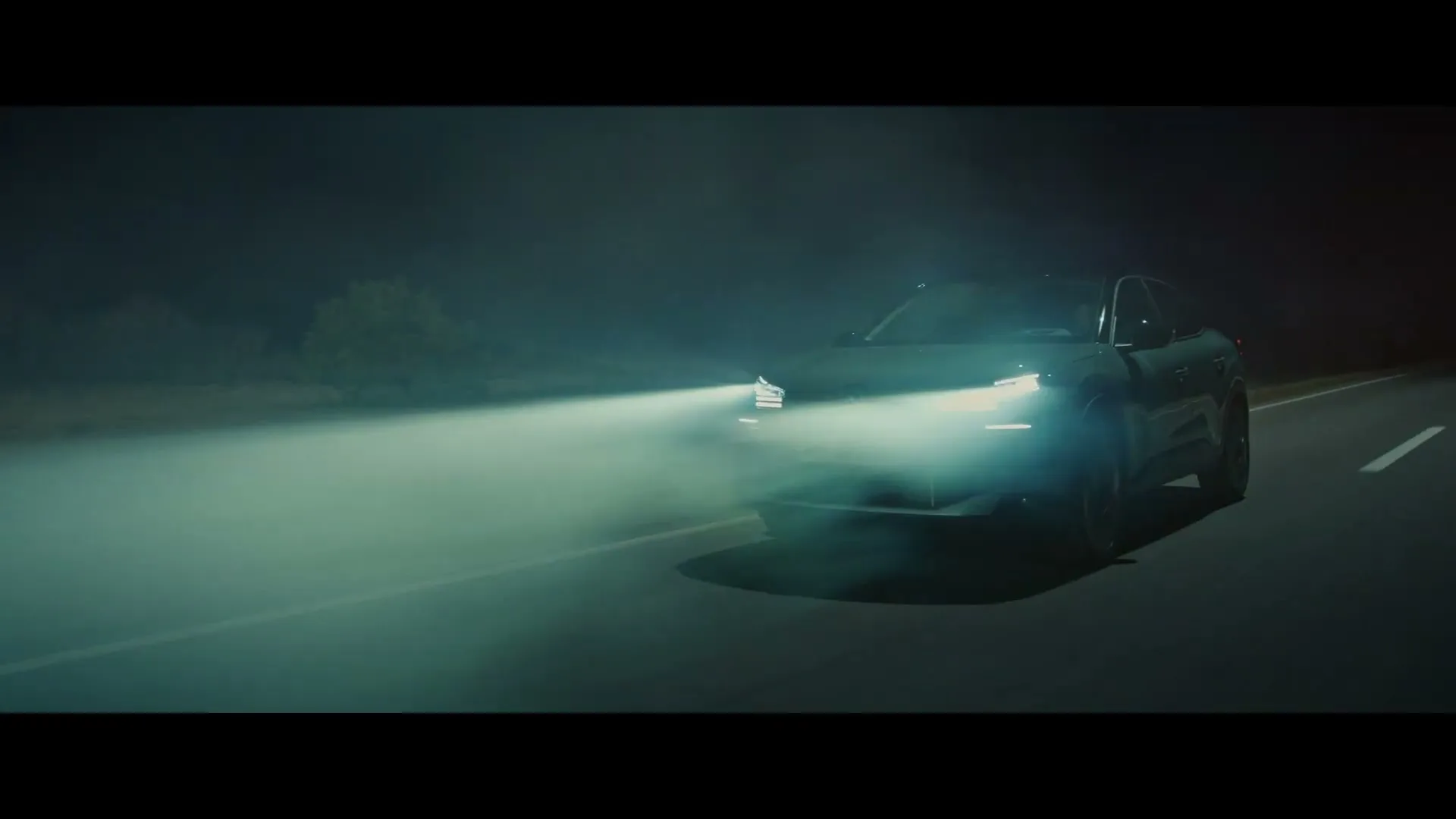 A teal Citroen car drives on a dark road at night, its headlights illuminating the misty air.