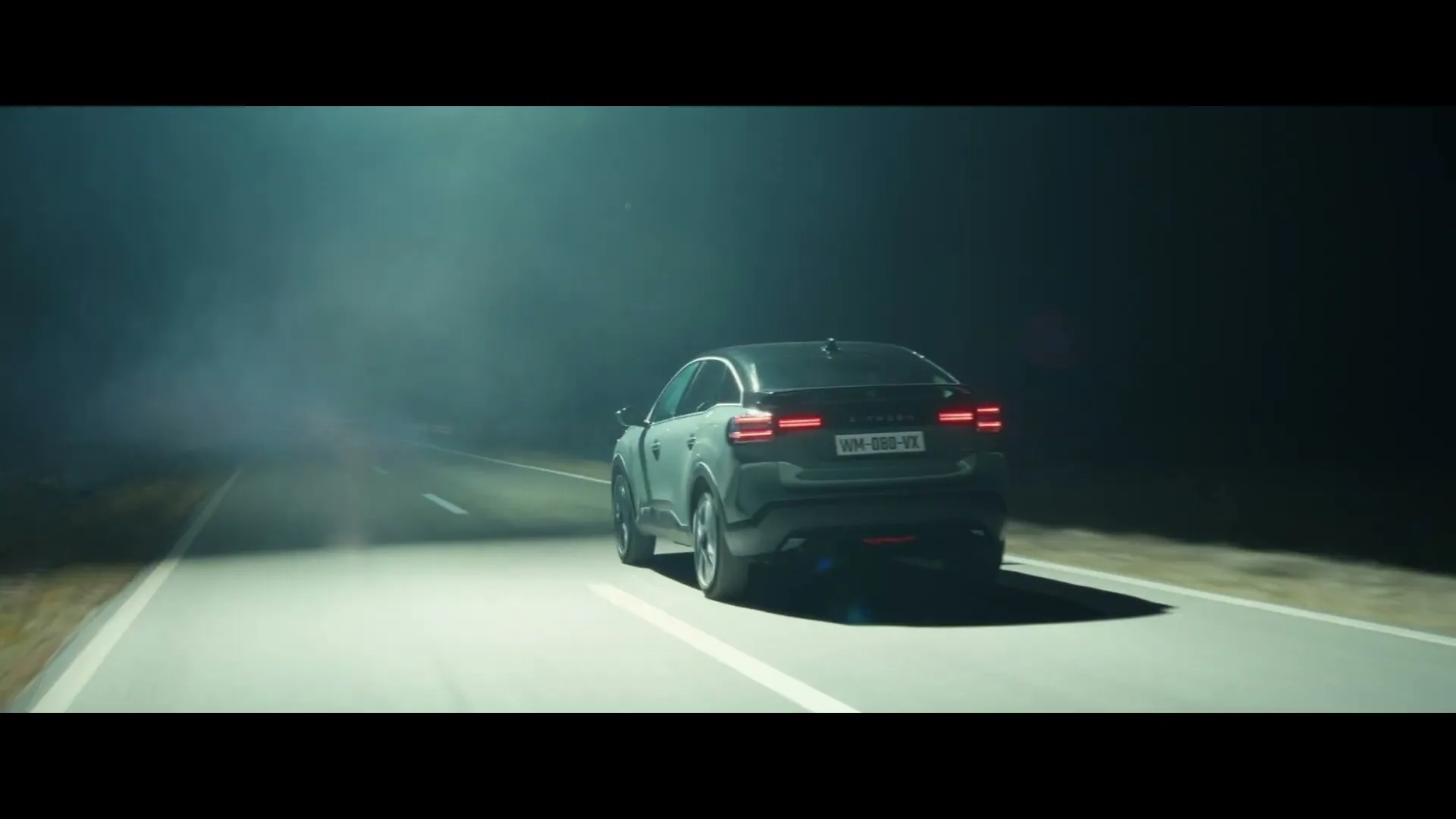 A dark grey Citroen car drives away from the camera on a dark, foggy road at night, with its taillights glowing red.