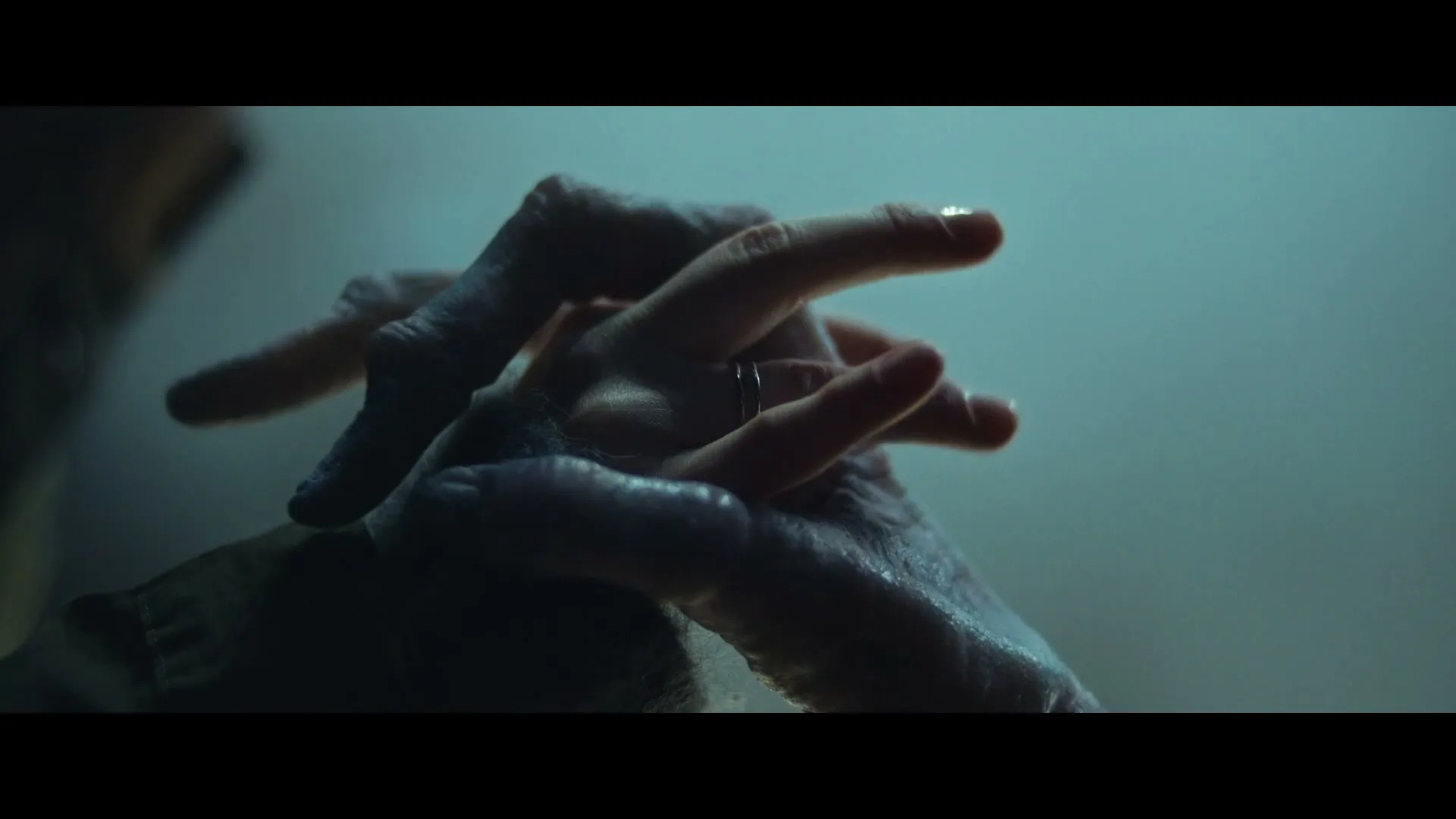 Two hands, one old and wrinkled, the other young and smooth with a ring, gently touch in a dimly lit, mysterious setting.