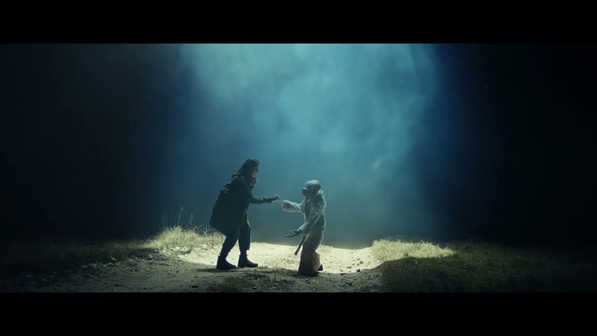 A woman in a dark coat and an alien creature in a light robe hold hands in a spotlight on a dirt path at night.