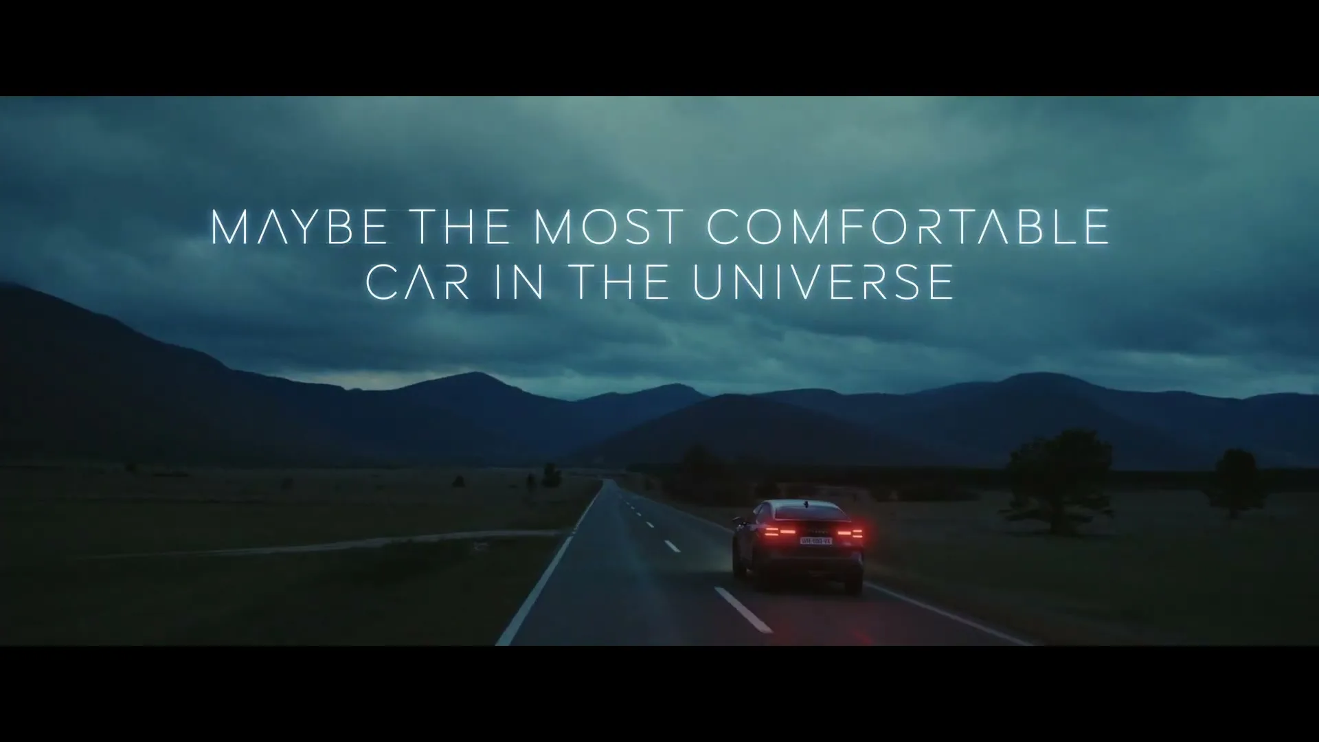 A dark car with glowing red taillights drives away on a long, empty road through a valley with mountains under a cloudy sky at dusk.