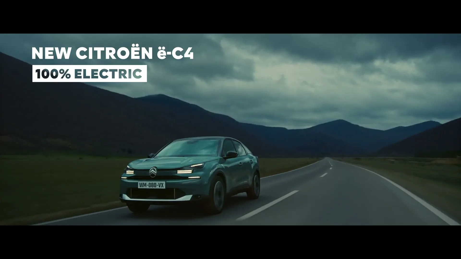 A green Citroen e-C4 drives on a winding road through a valley, with a UFO hovering above and mountains in the background under a cloudy sky.