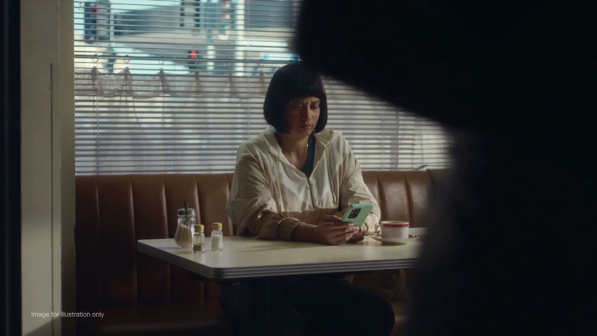 A woman with a bob haircut sits in a diner booth, looking at her phone with a concerned expression, as a waiter walks by.