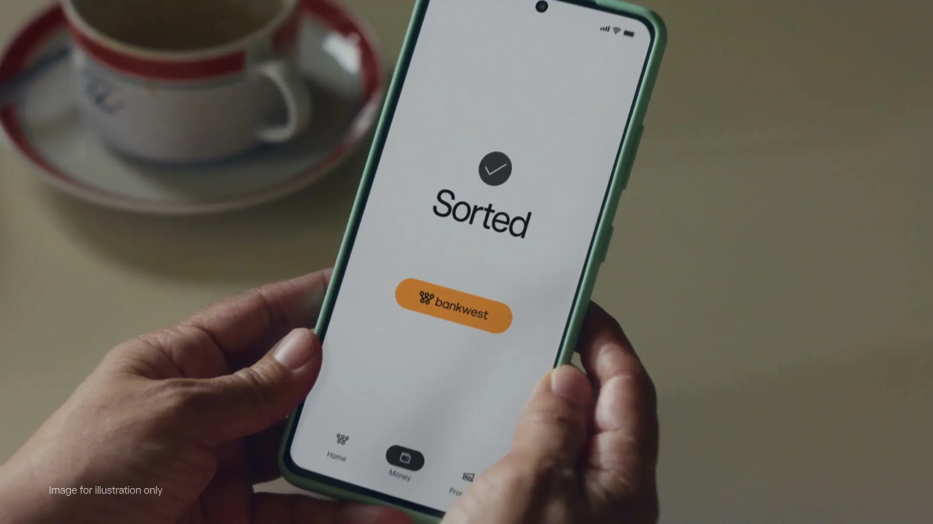 A person's hands hold a smartphone displaying the Bankwest app with "Sorted" and the Bankwest logo, next to a coffee cup on a table.