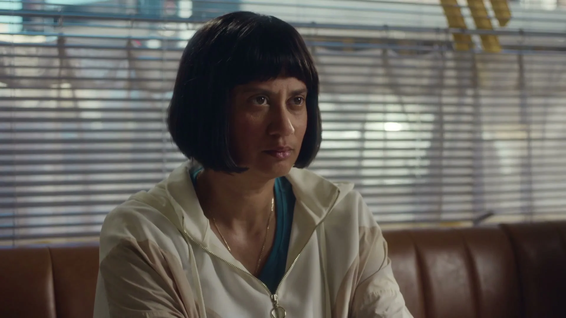 A woman with a bob haircut and a white jacket sits on a brown booth, looking intently to the right in an interior setting.