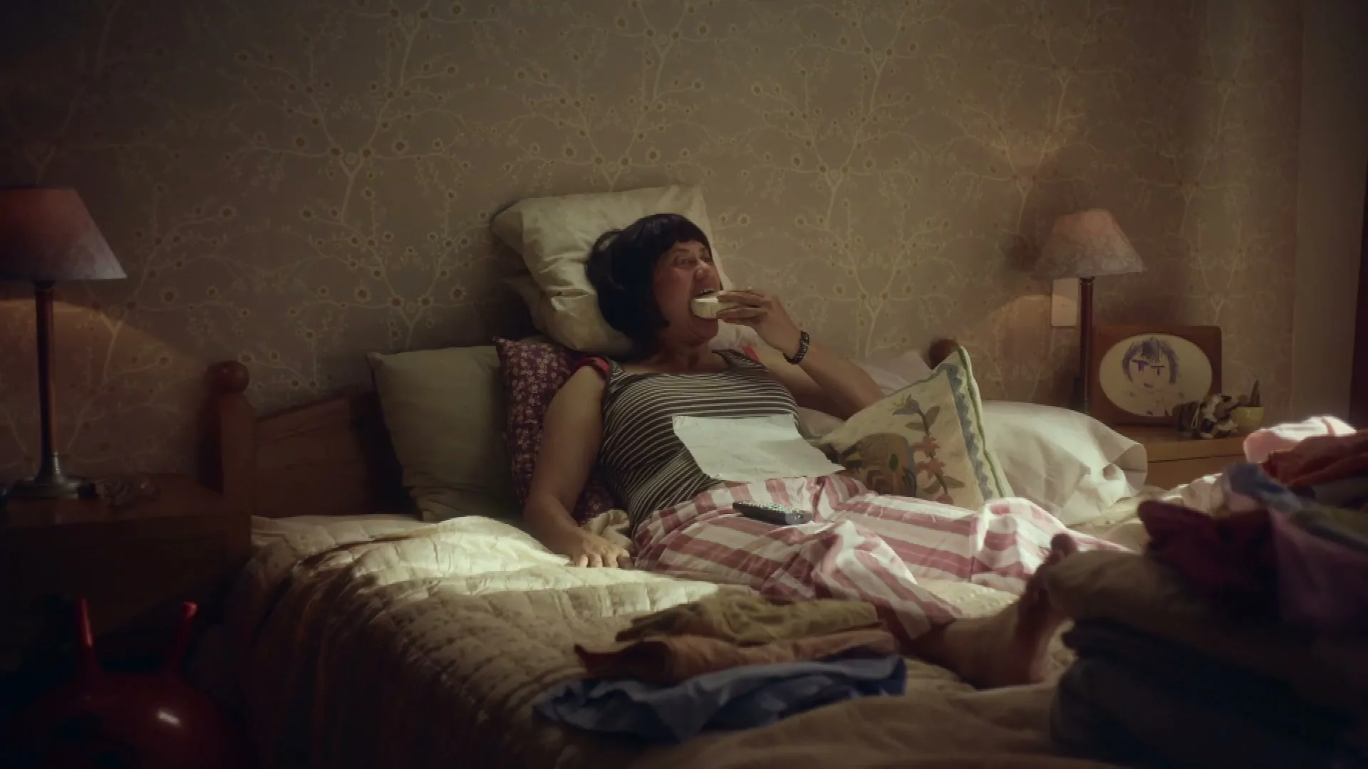 A woman with dark hair and a striped top lies in bed, eating a snack while watching TV in a dimly lit bedroom.