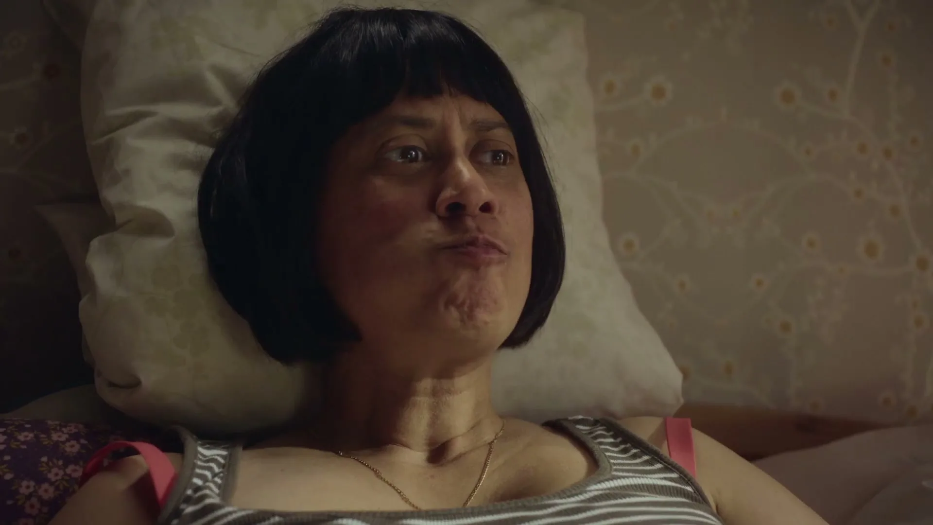 A woman with a bob haircut lies in bed, eating a piece of cheese, with a patterned wallpaper in the background.