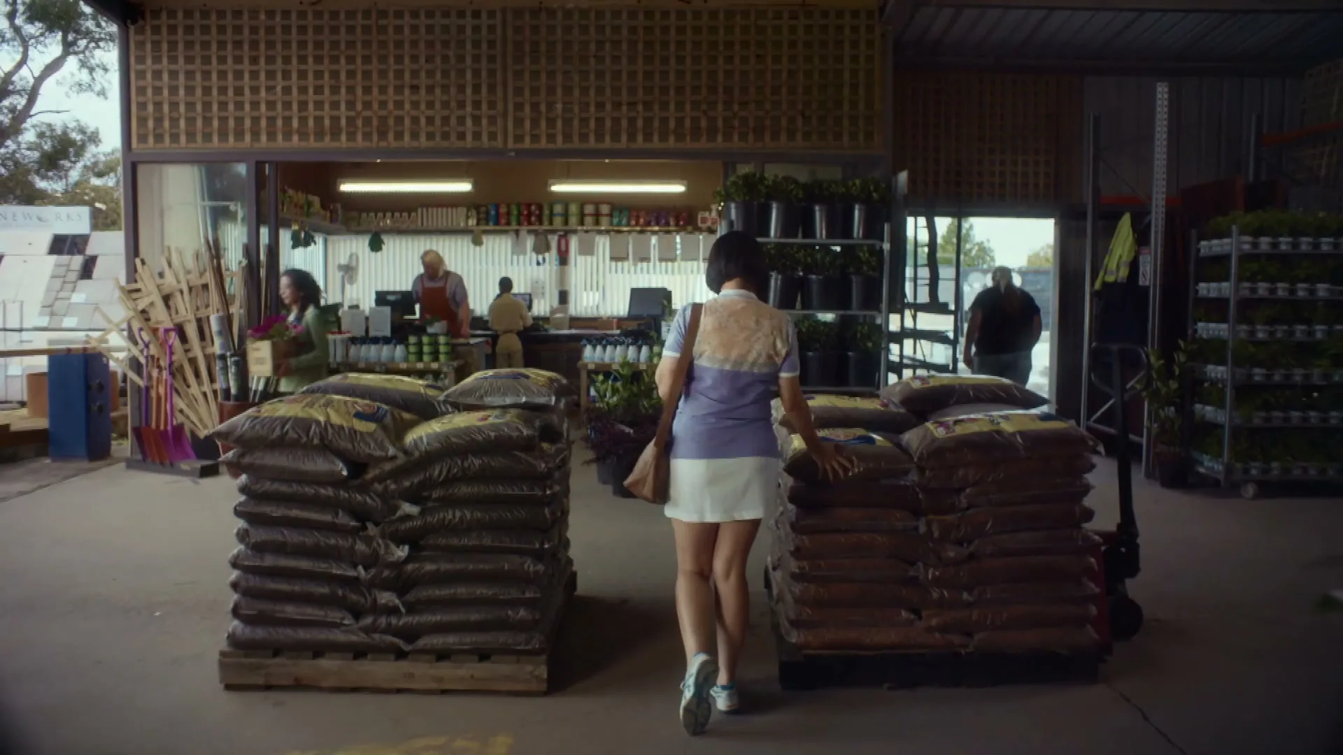 A young woman with short dark hair, wearing a polo shirt and white skirt, walks through a garden center past stacks of soil bags.