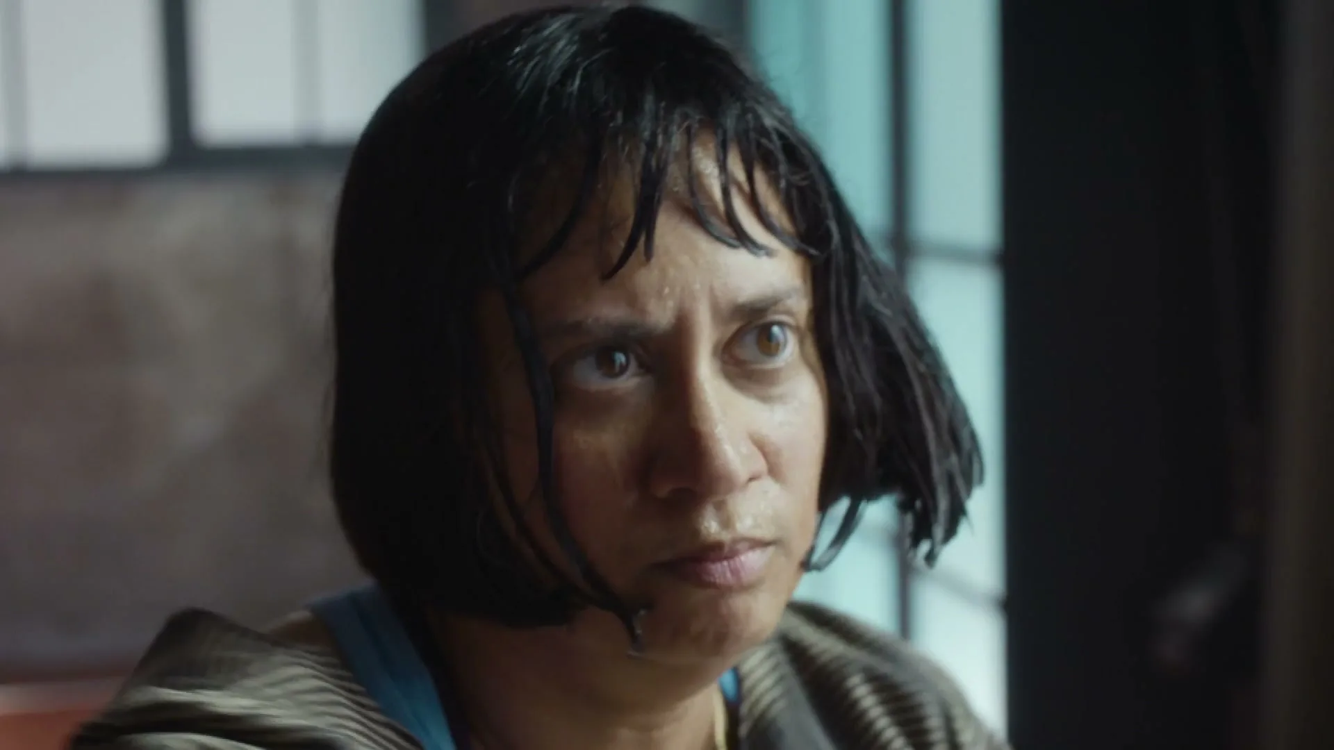 A woman with short, dark hair and a striped top looks intently at something off-screen, her expression serious, in an interior setting.