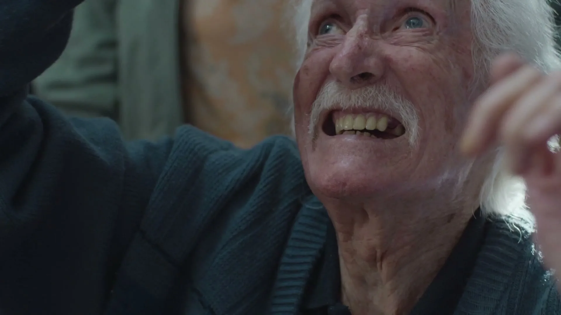 An elderly man with white hair and a mustache looks up with a surprised expression, raising his hands in an interior setting.
