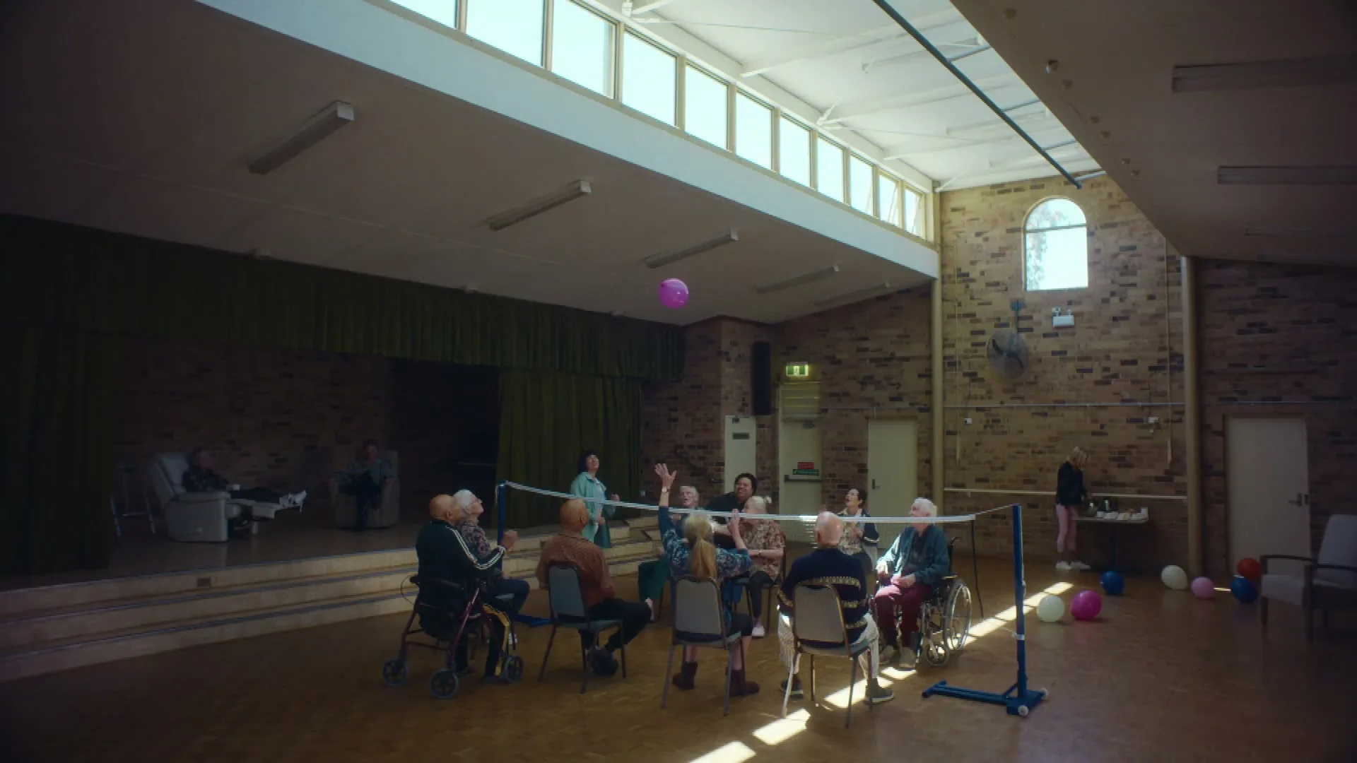 A group of elderly people play balloon volleyball in a community hall, with one woman hitting the balloon over the net.