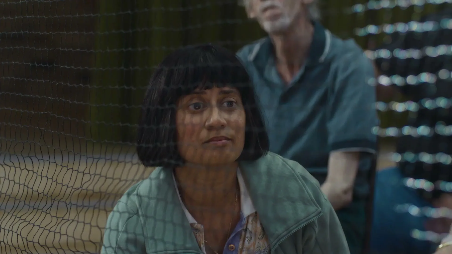 A woman with a bob haircut and a green jacket looks up, then forward, through a chain-link fence at a baseball game.