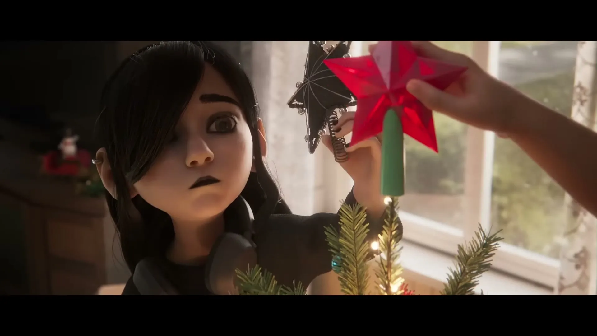 A young animated girl with black hair and dark clothes tries to place a black star on a Christmas tree, but an adult hand replaces it with a red star in a cozy living room.